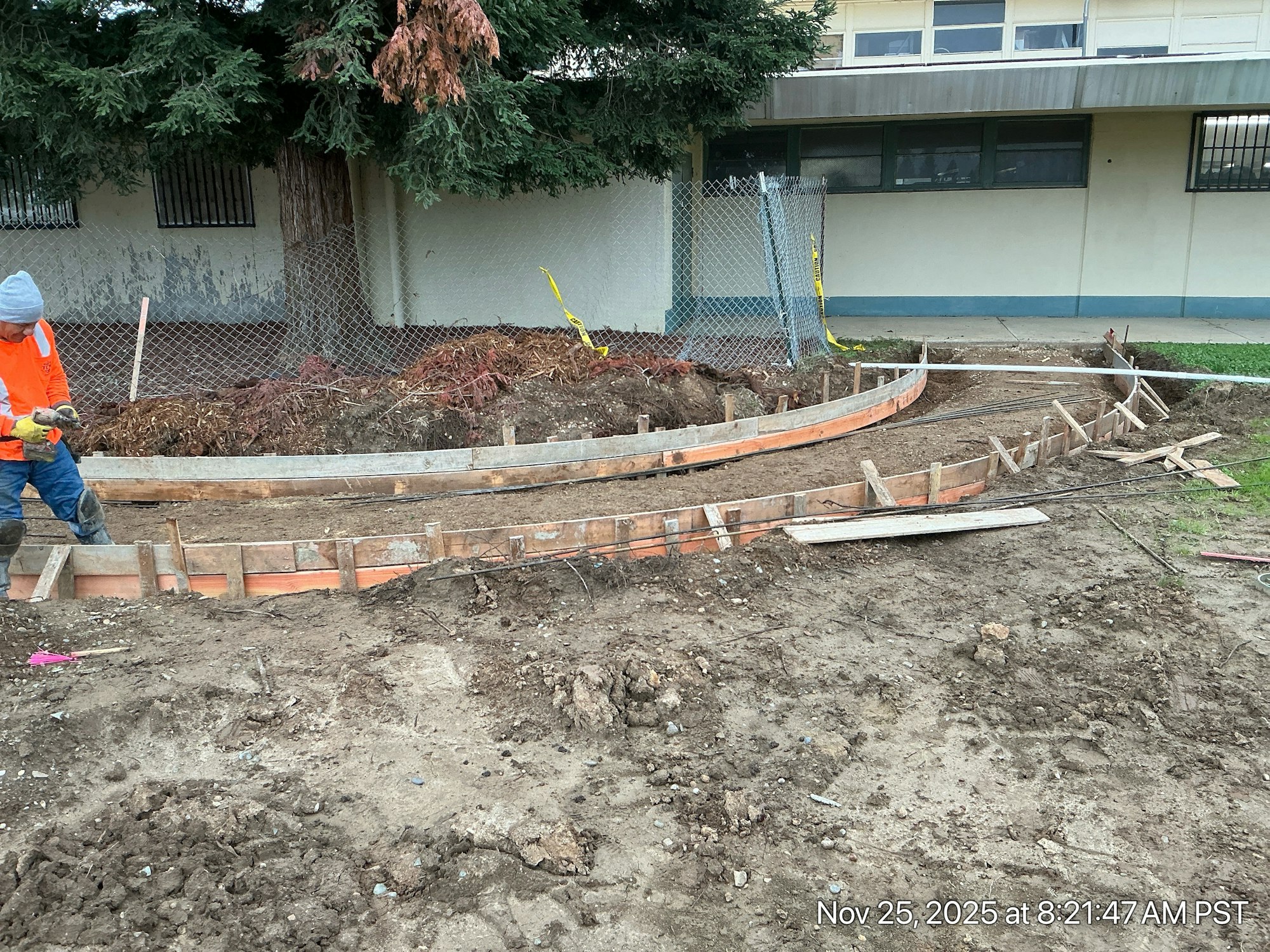 A construction site with a worker in an orange vest preparing forms for a concrete slab, set against a school background.