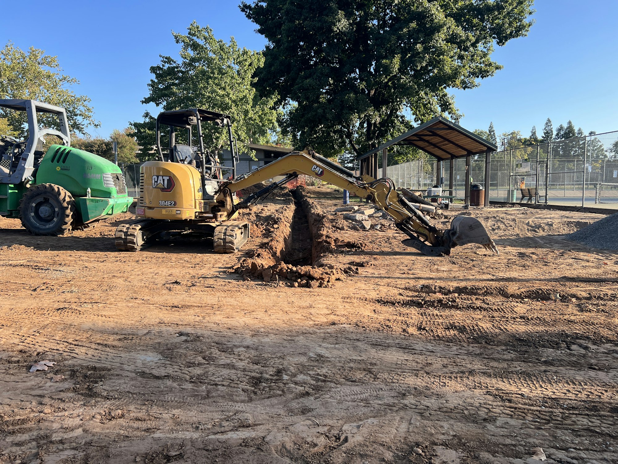 Construction site with a mini excavator digging and a green machinery vehicle nearby, surrounded by trees and a fenced area.