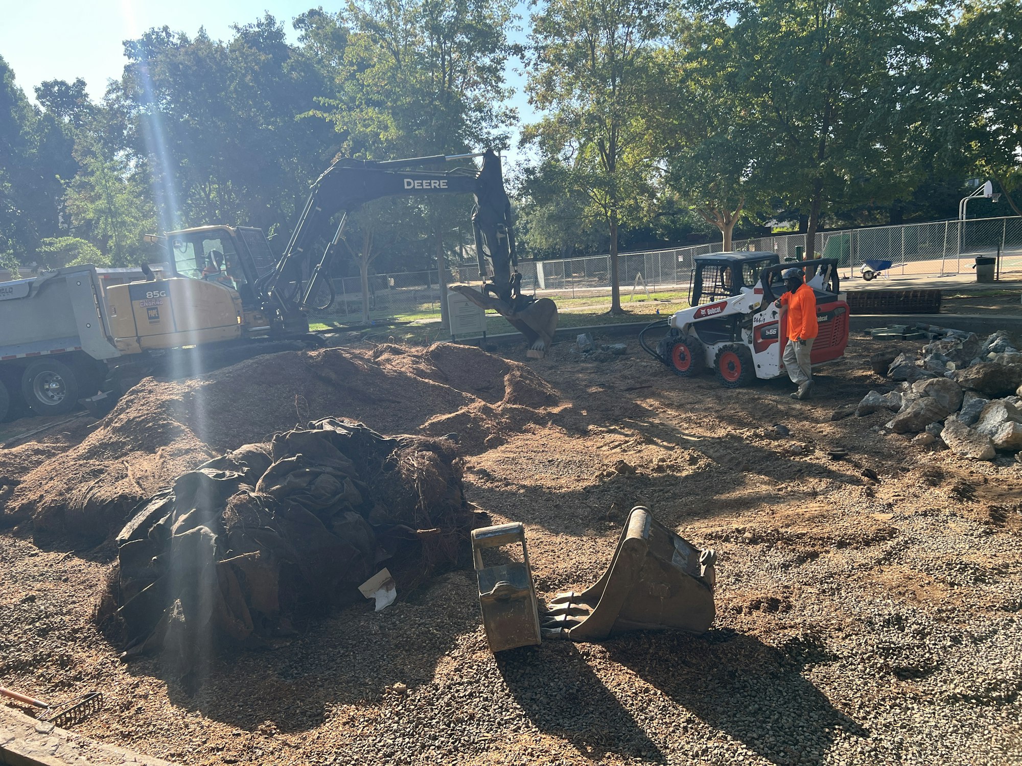 A construction scene with an excavator and skid steer loader, workers, gravel piles, and trees in the background.