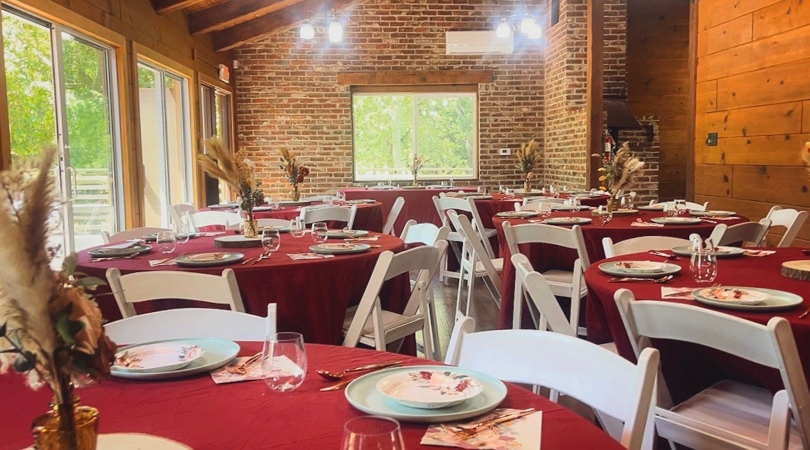Rustic dining area with round tables, white chairs, red tablecloths, and decorative centerpieces. Brick walls and large windows.