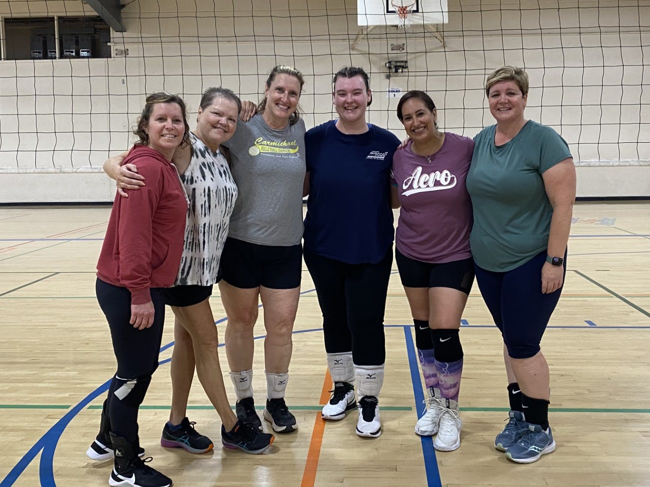 A group of six women poses together in a gym with a volleyball net in the background, smiling and dressed in athletic wear.