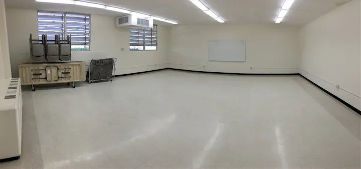 The image shows a sparsely furnished room with some stacked chairs, a folding table, and a whiteboard on the wall.