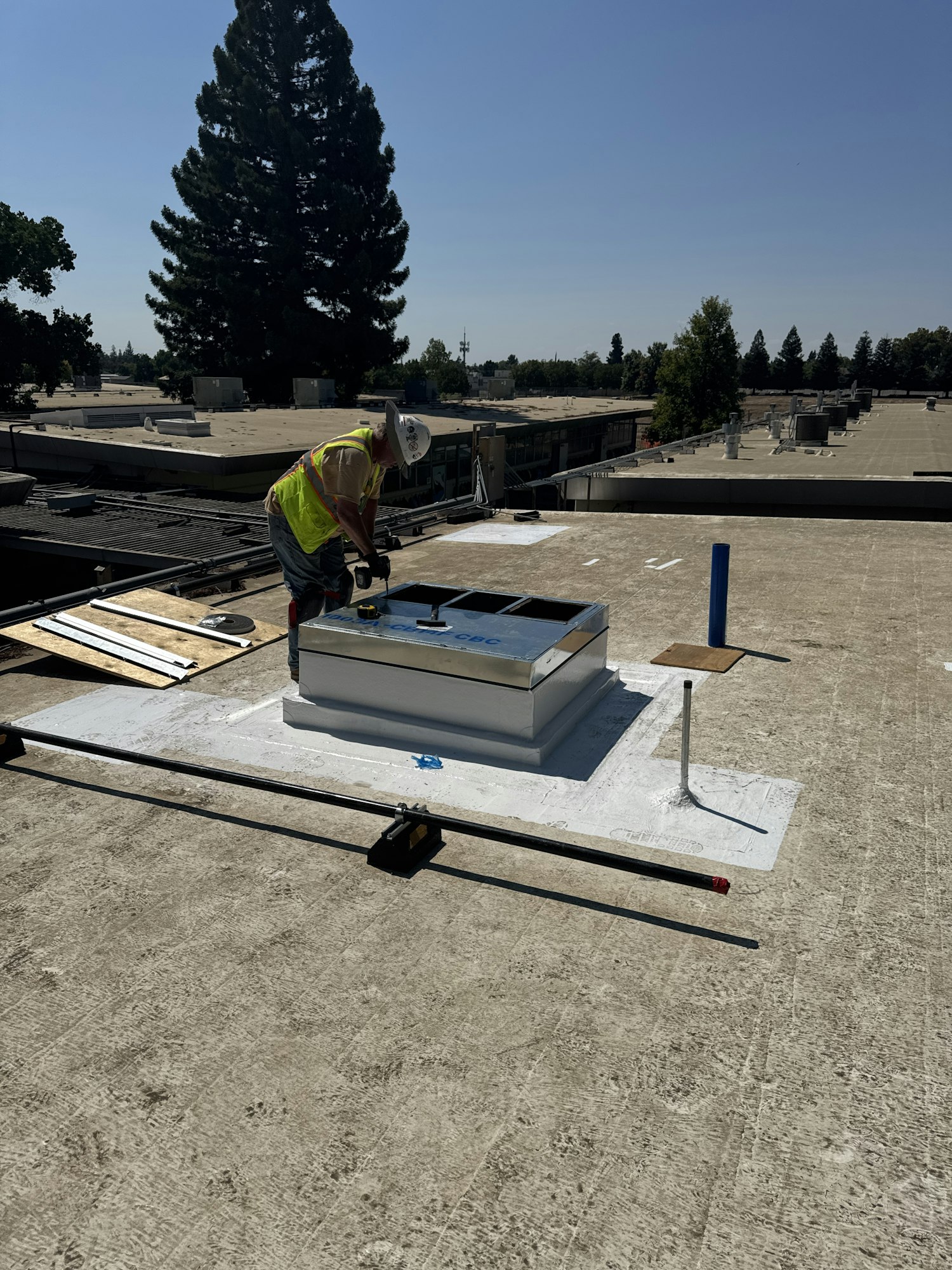 A construction worker in safety gear working on a rooftop with tools and materials under a clear sky.