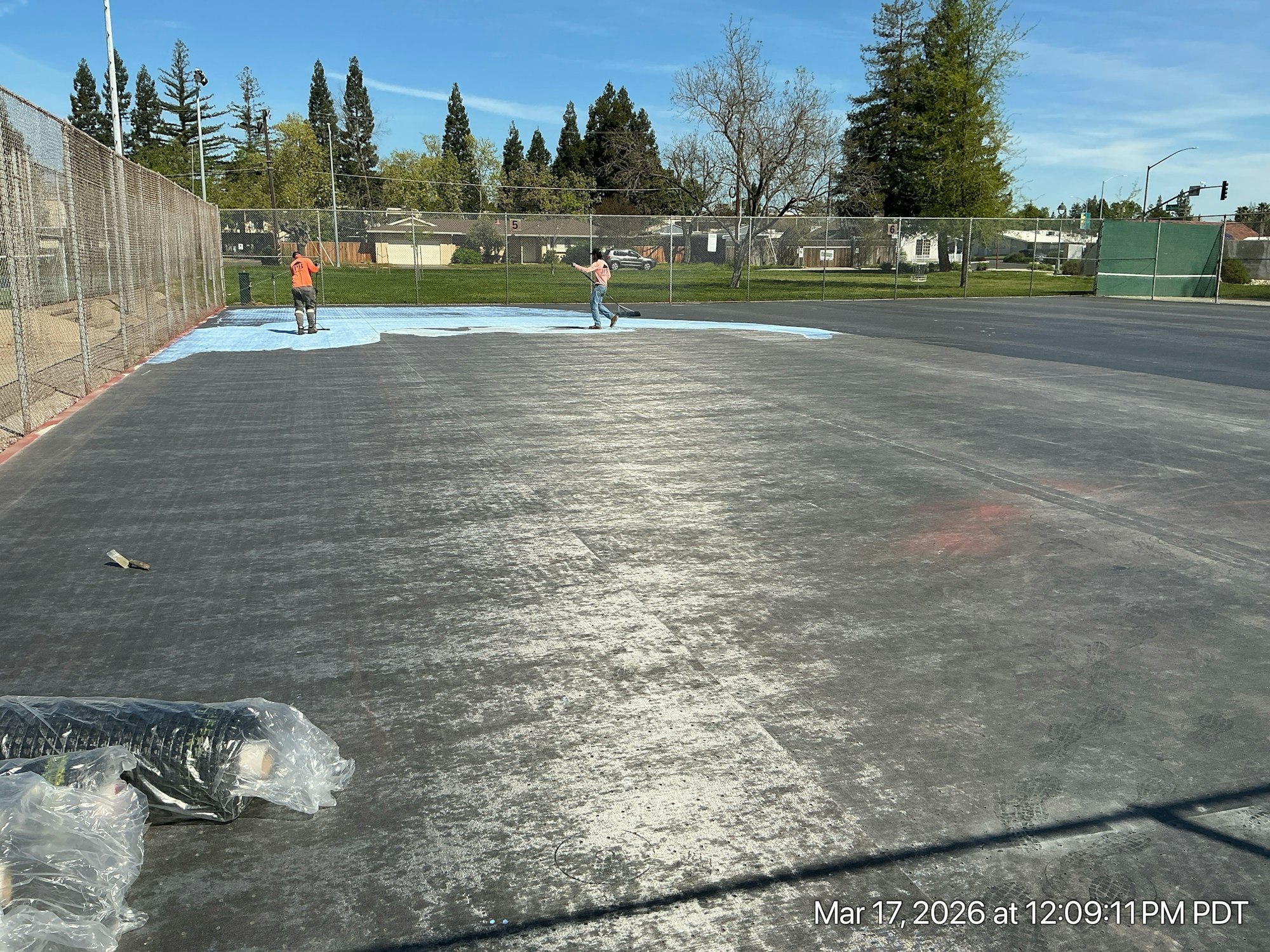 Workers are applying a surface cover on an outdoor court or field, surrounded by a fence and green spaces in the background.