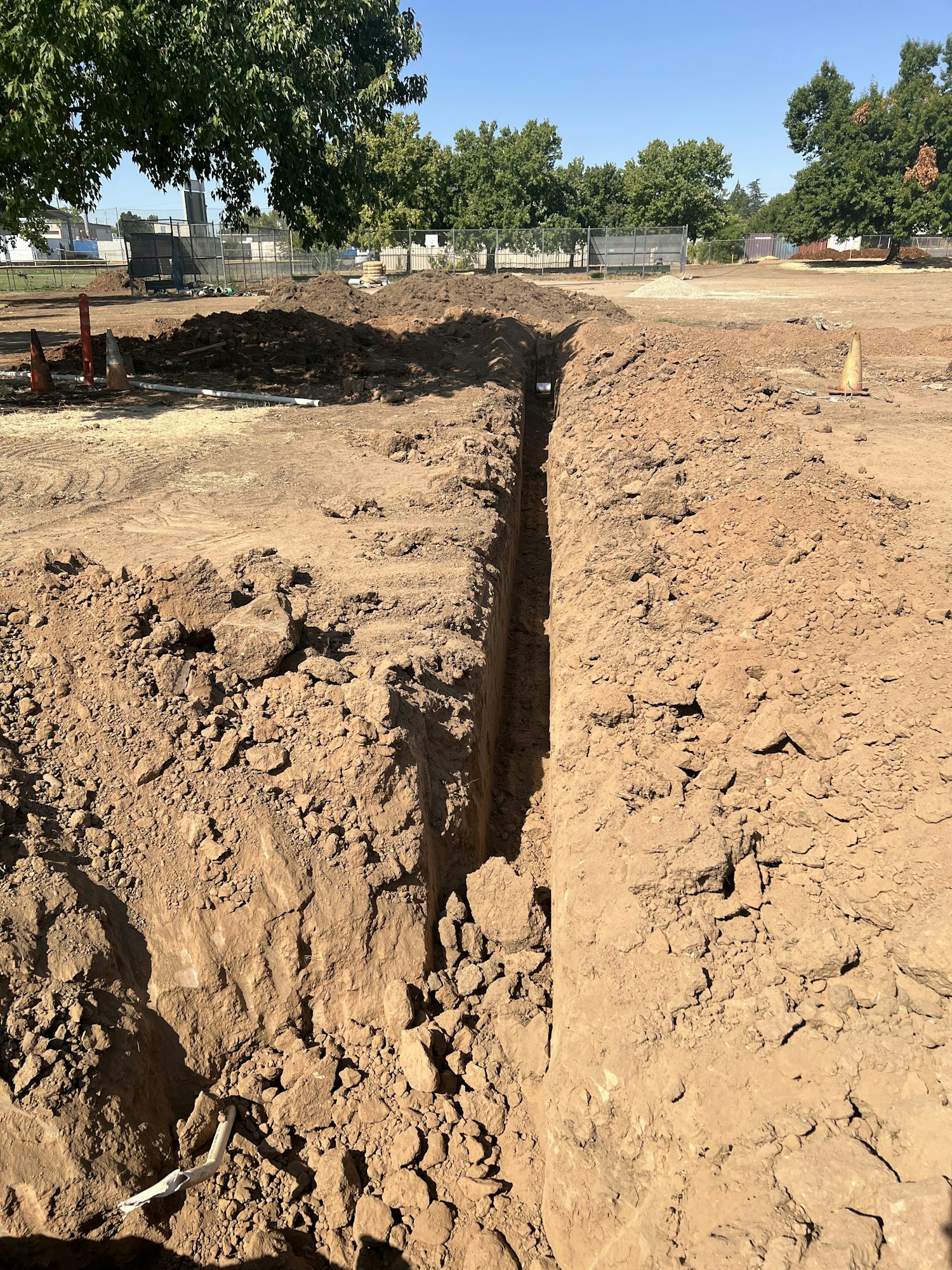 A trench dug in dirt at a construction site, surrounded by trees and fencing.