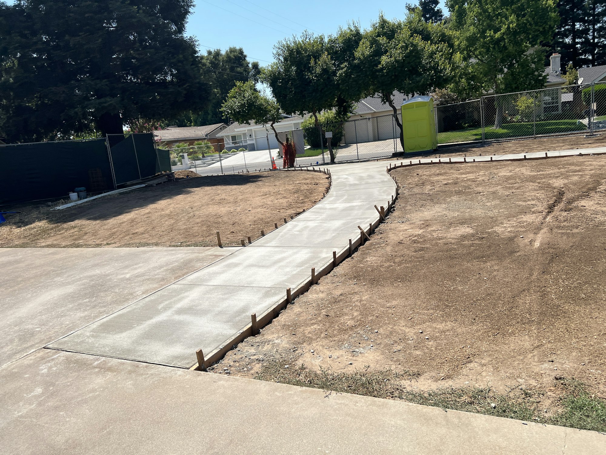 A newly paved concrete path winding through dirt and grass, with a fence and portable toilet in the background.