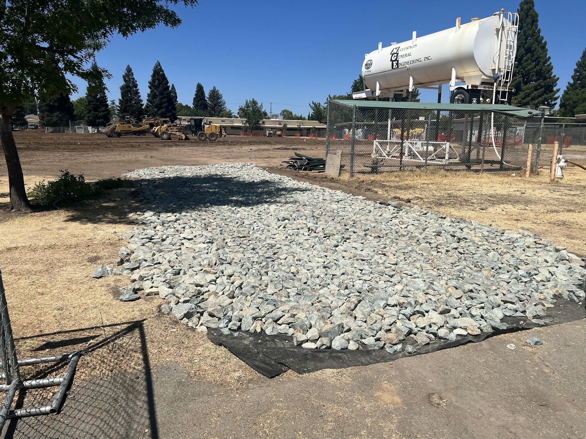 Construction site with gravel, machinery, a fenced area, and a large industrial tank in the background.