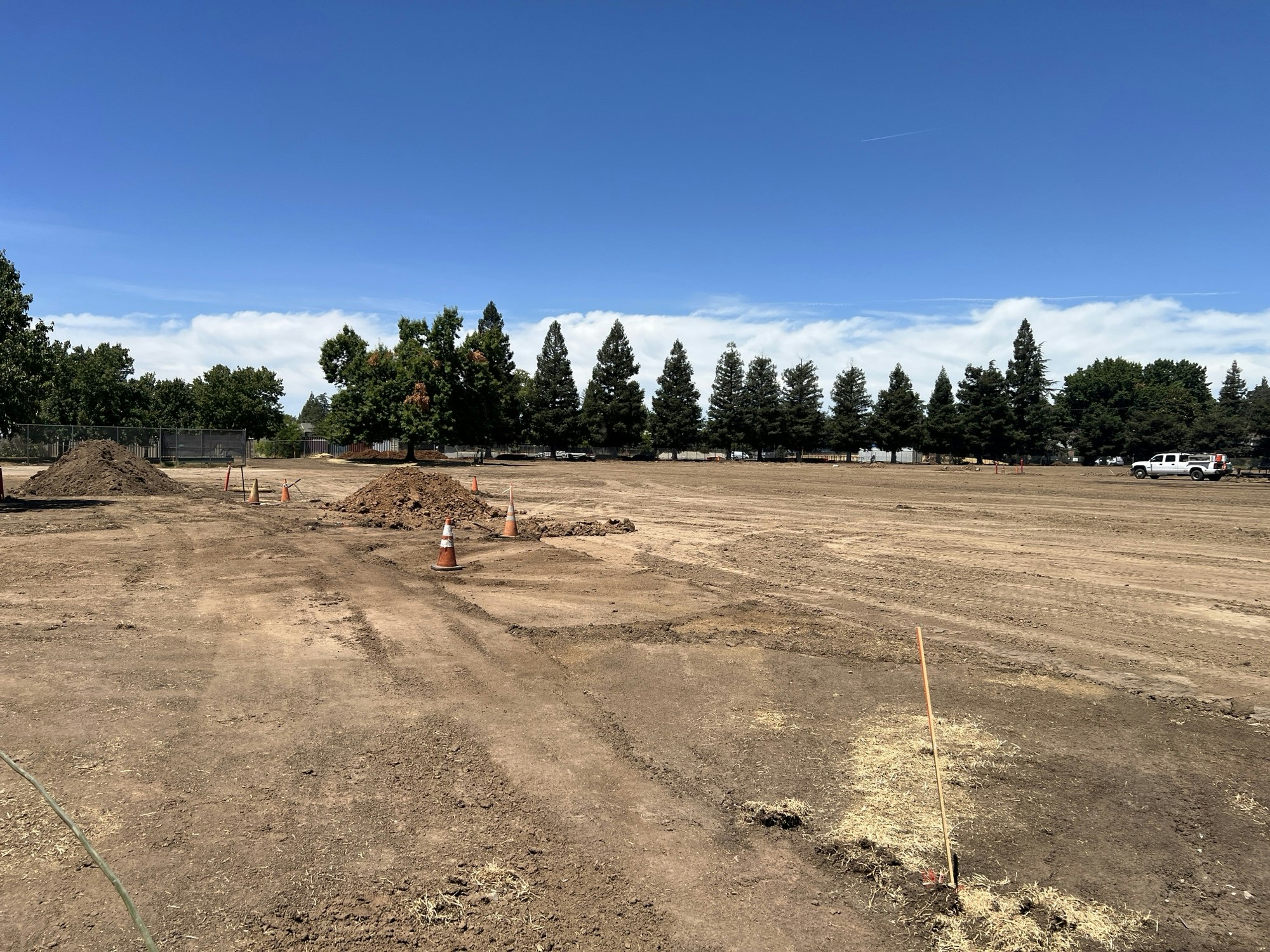 Construction site with dirt piles, traffic cones, and a truck in the background. Trees line the horizon under a clear blue sky.