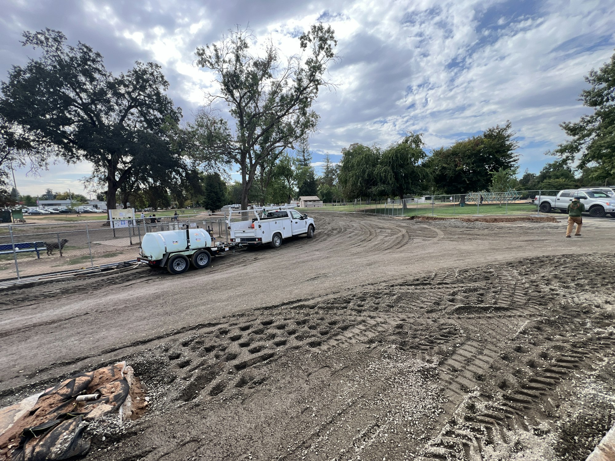 Construction site with trucks, tire tracks on dirt, trees, and cloudy sky.