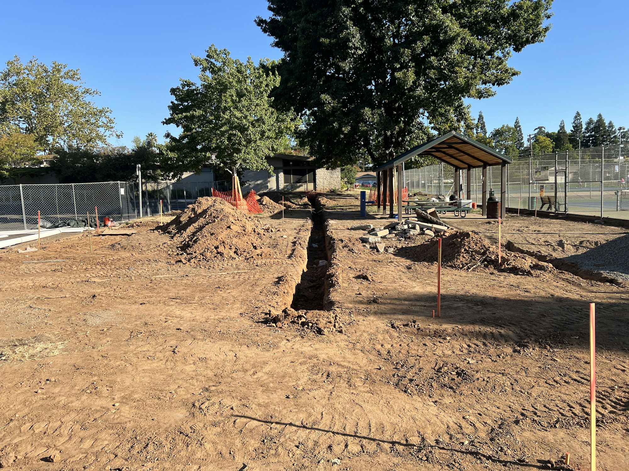 Construction site with dug trenches, piles of dirt, a tree, a small shelter, and a fence, set in a sunny area.