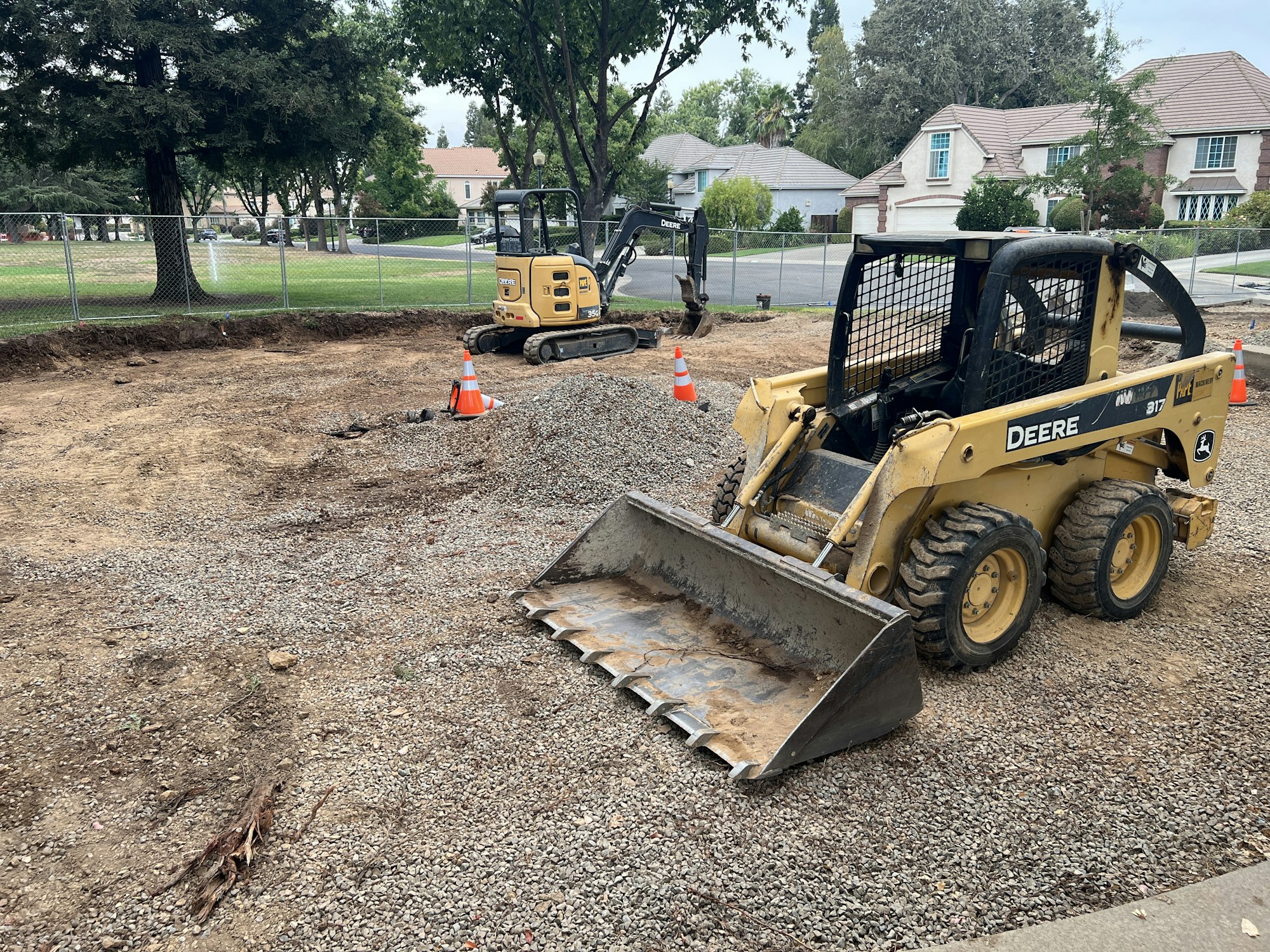 Construction site with machinery, including a skid steer loader and an excavator, surrounded by traffic cones and a fence.