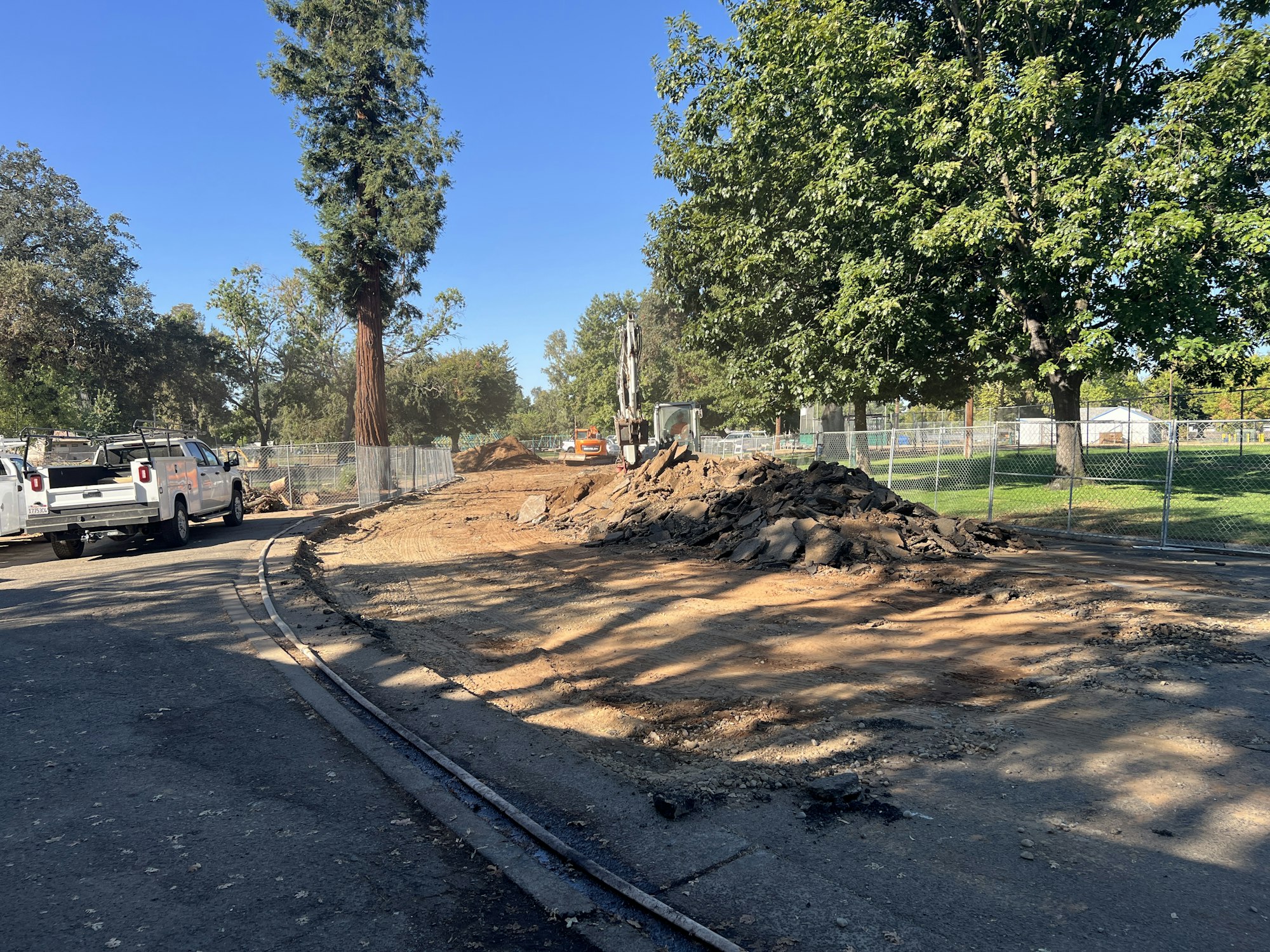 Construction site with dirt piles, trucks, fencing, and trees along the edge of a park area.