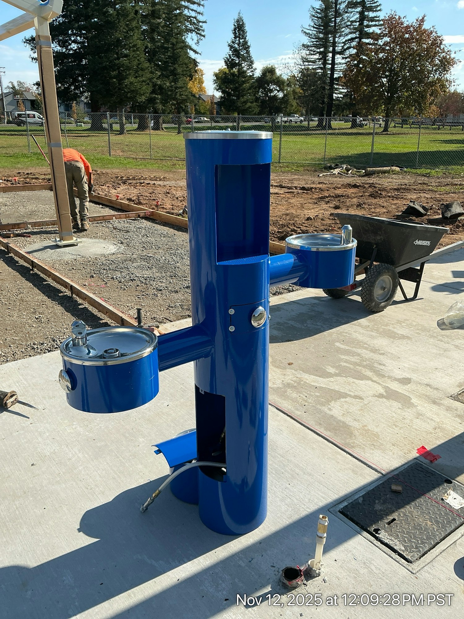 A blue drinking fountain, part of a construction site, with trees and a worker nearby.