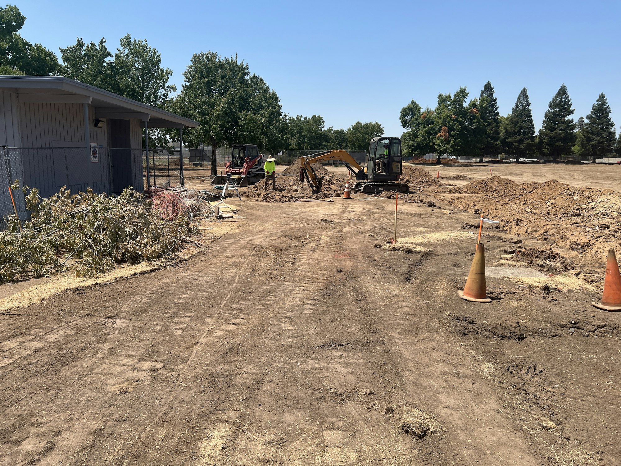 Construction site with a small excavator, dirt piles, orange cones, and some trees in the background.