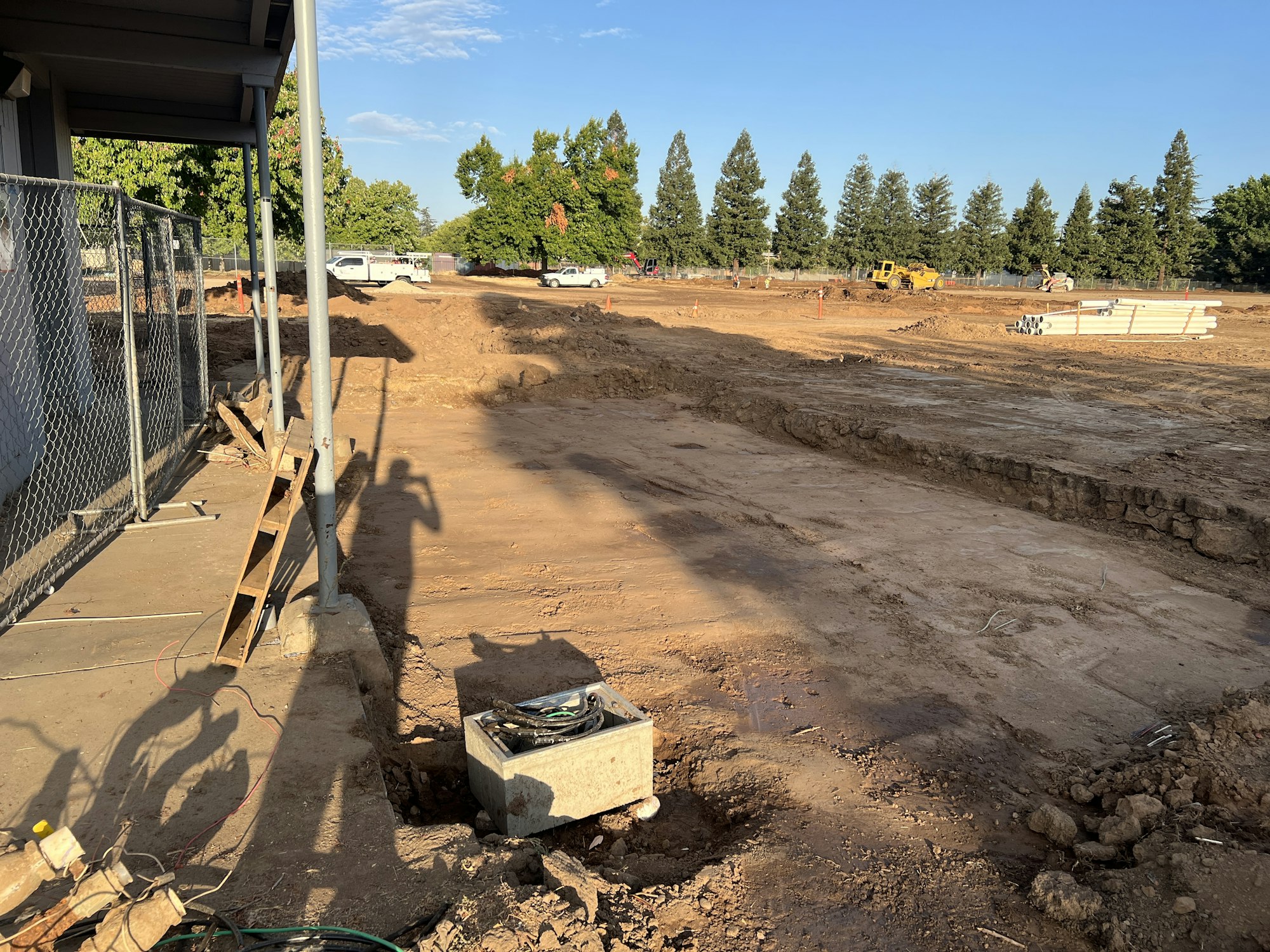 Construction site with dug-out ground, metal pipes, fence, and trees in the background. Shadows and sunlight visible.