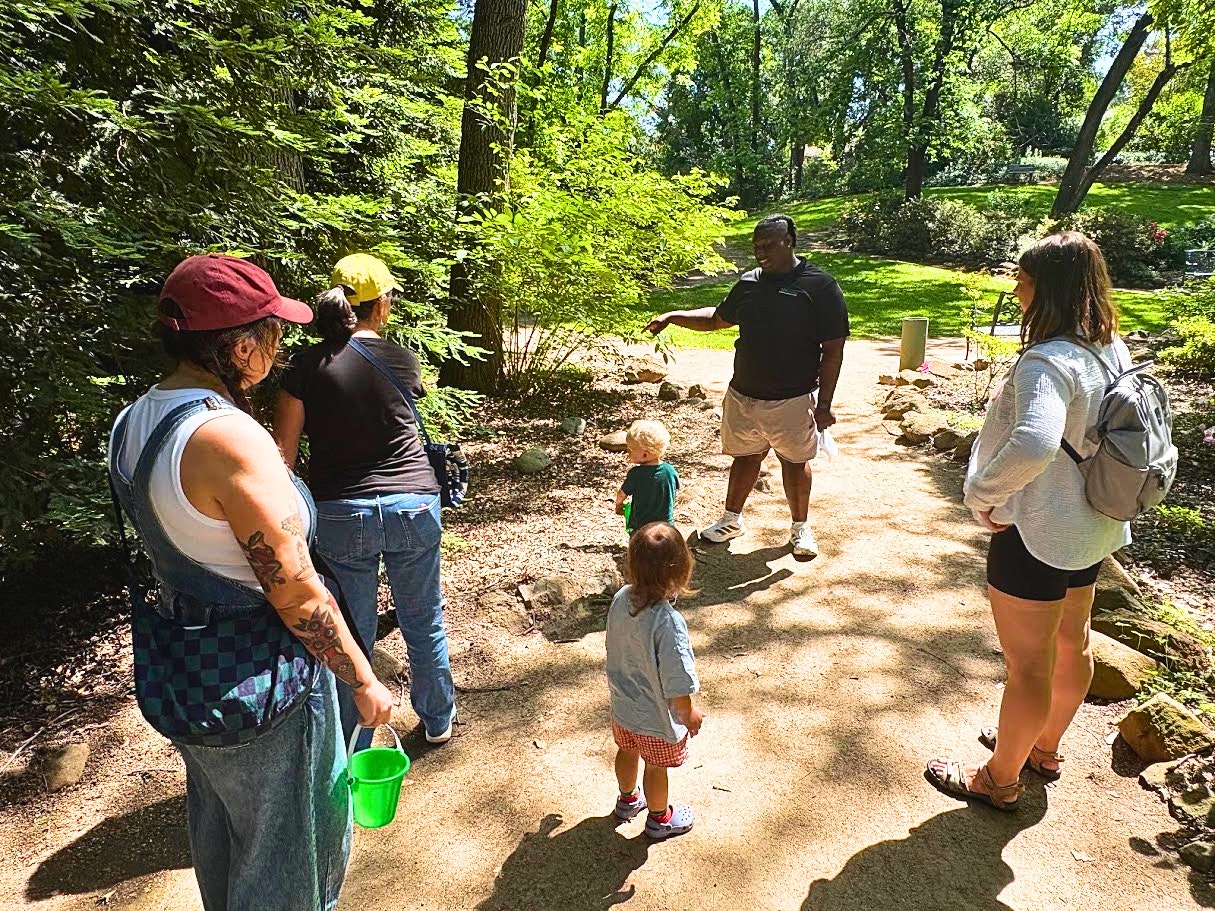 A group of adults and children gathered in a lush outdoor setting, with one adult speaking to the group.