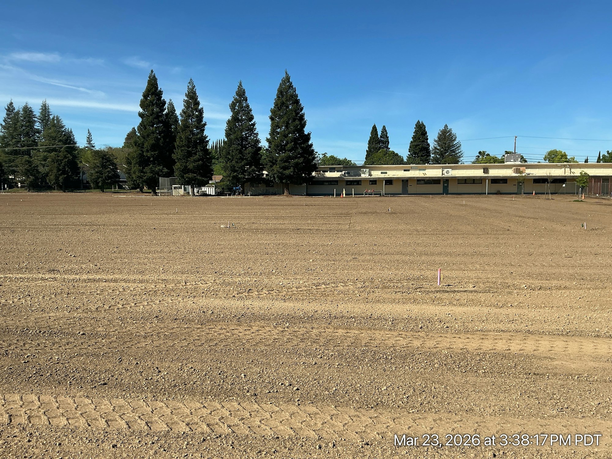 A cleared, flat dirt field with some markers and tall trees in the background, near a low building under a blue sky.