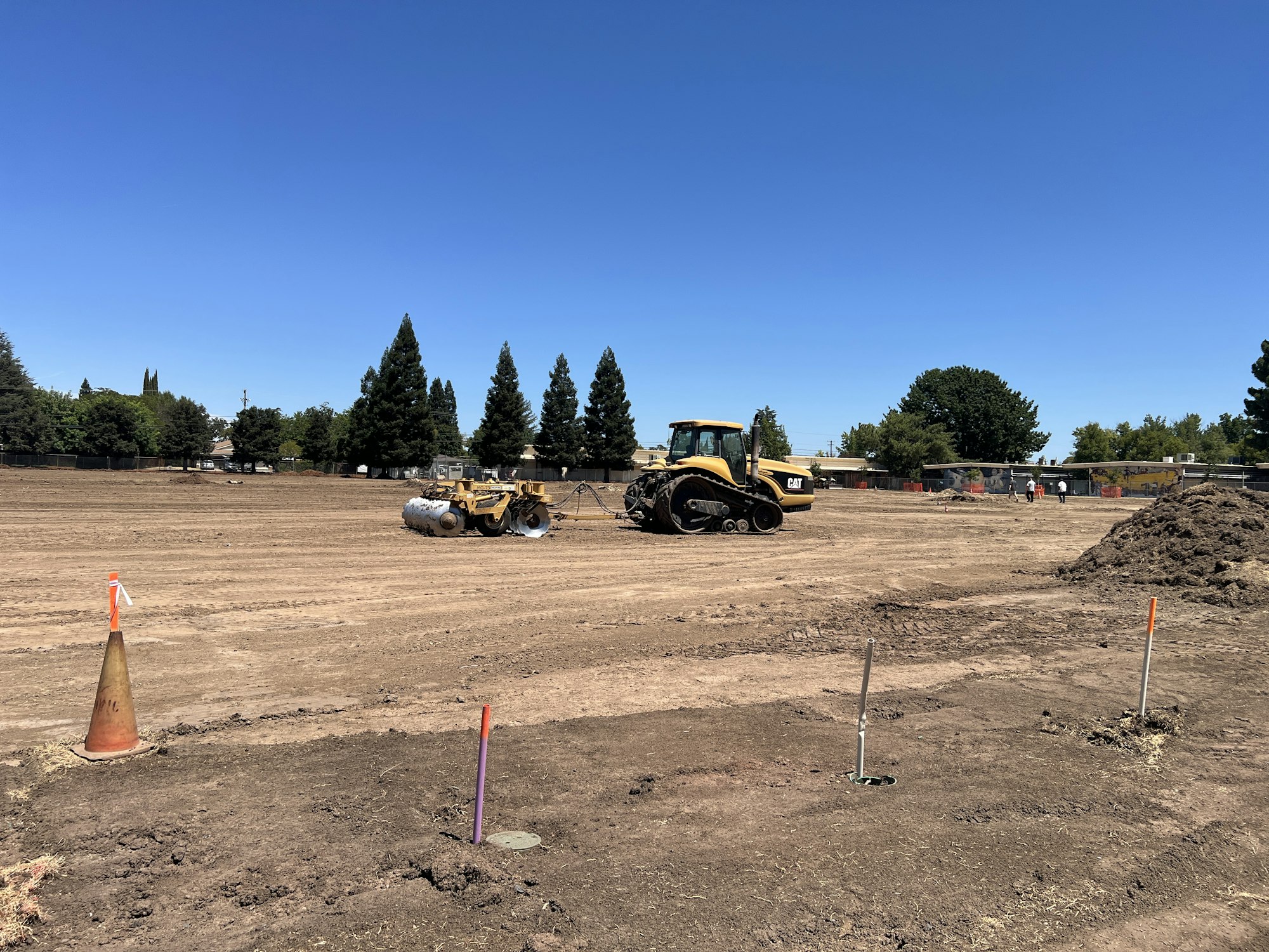 Construction site with a bulldozer, roller, cones, and poles on a dirt field under a clear blue sky.
