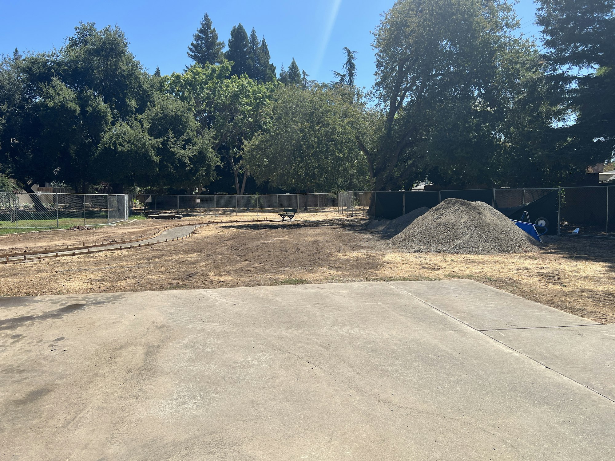 A yard with concrete, dirt, a gravel pile, a wheelbarrow, trees, and a fenced area.