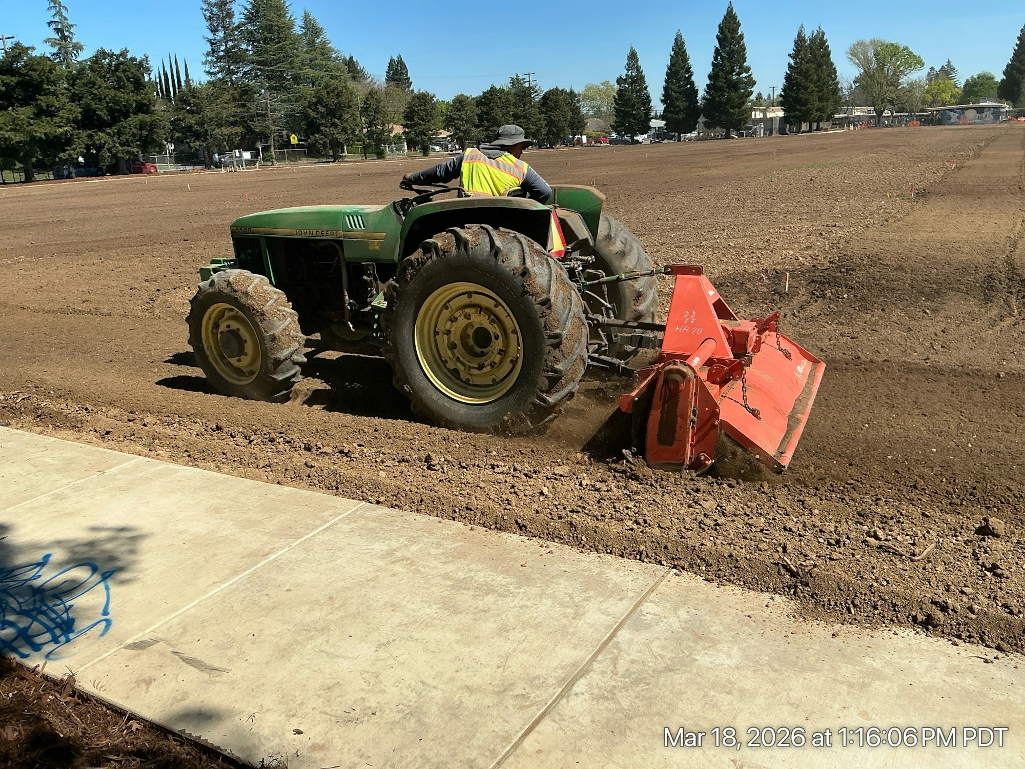 A tractor is plowing a dirt field under clear blue skies, with trees and a sidewalk in the background.