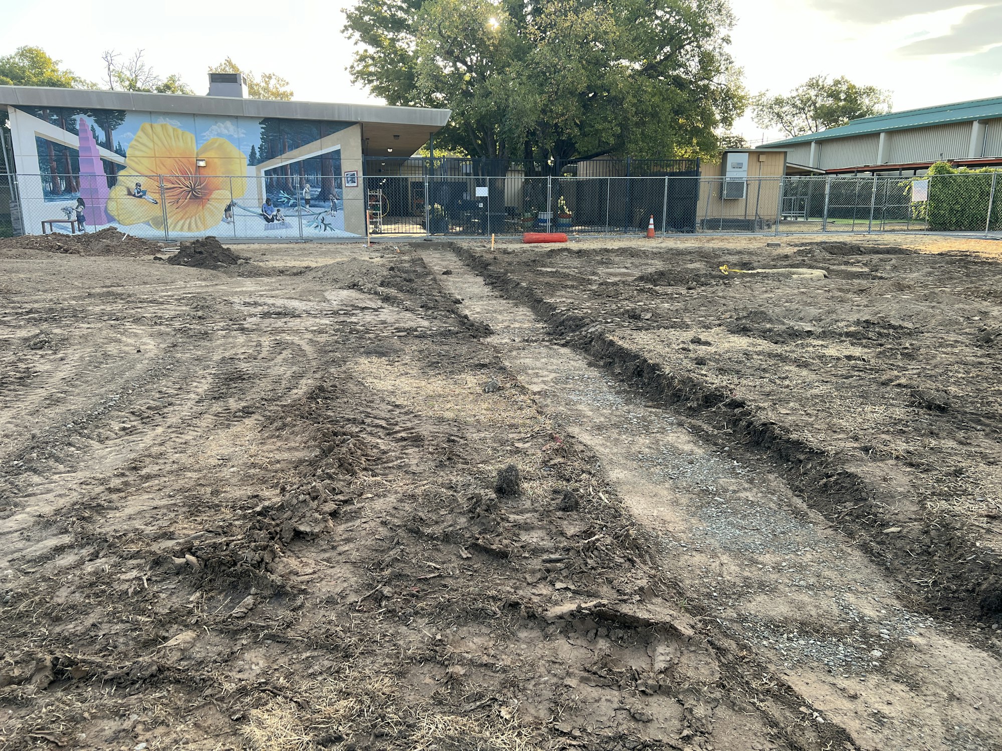 Dirt construction site with tracks, bordered by a building with a large mural and a chain-link fence.