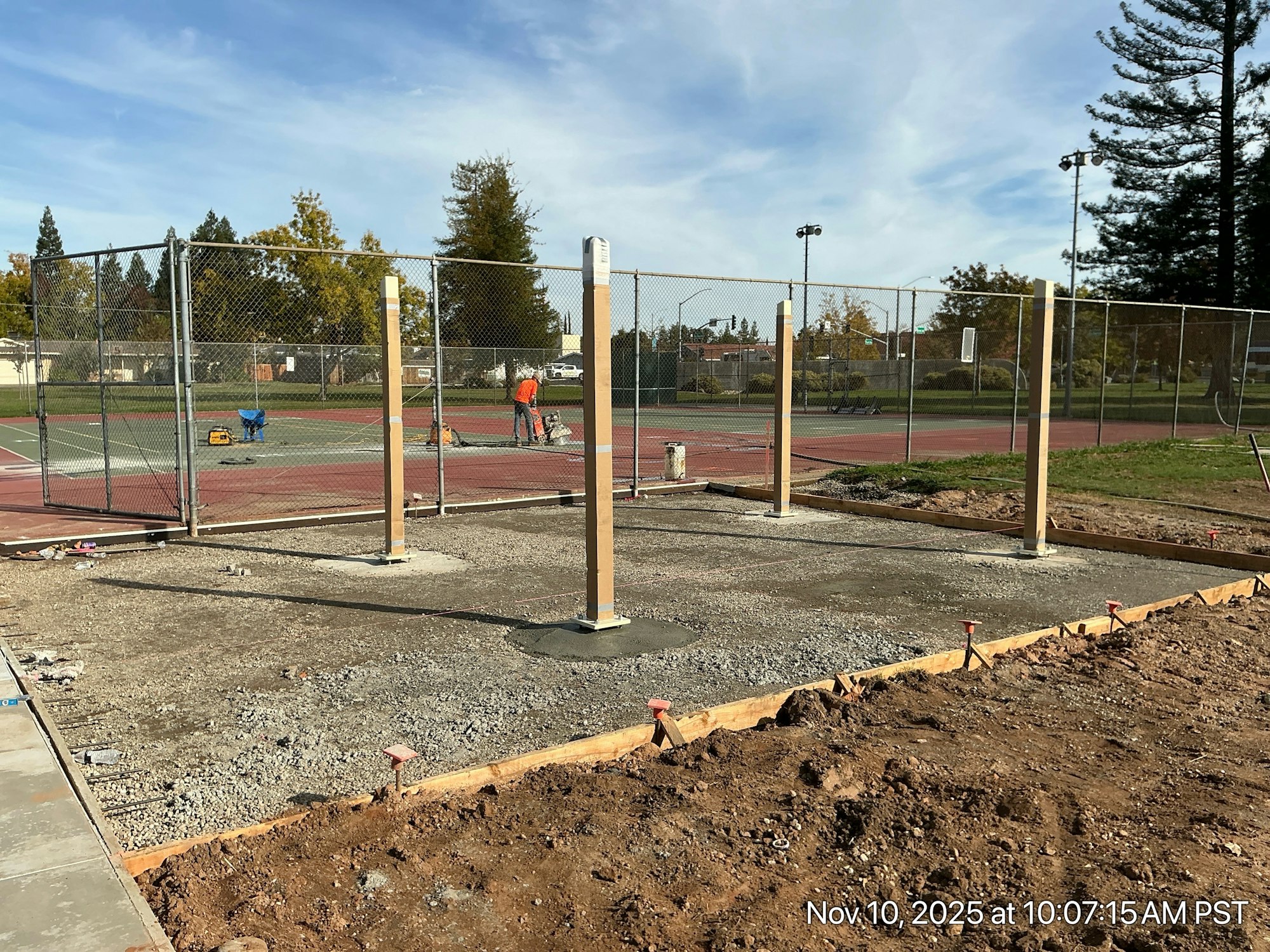 The image shows construction work on a site near tennis courts, with wooden posts and workers in action.