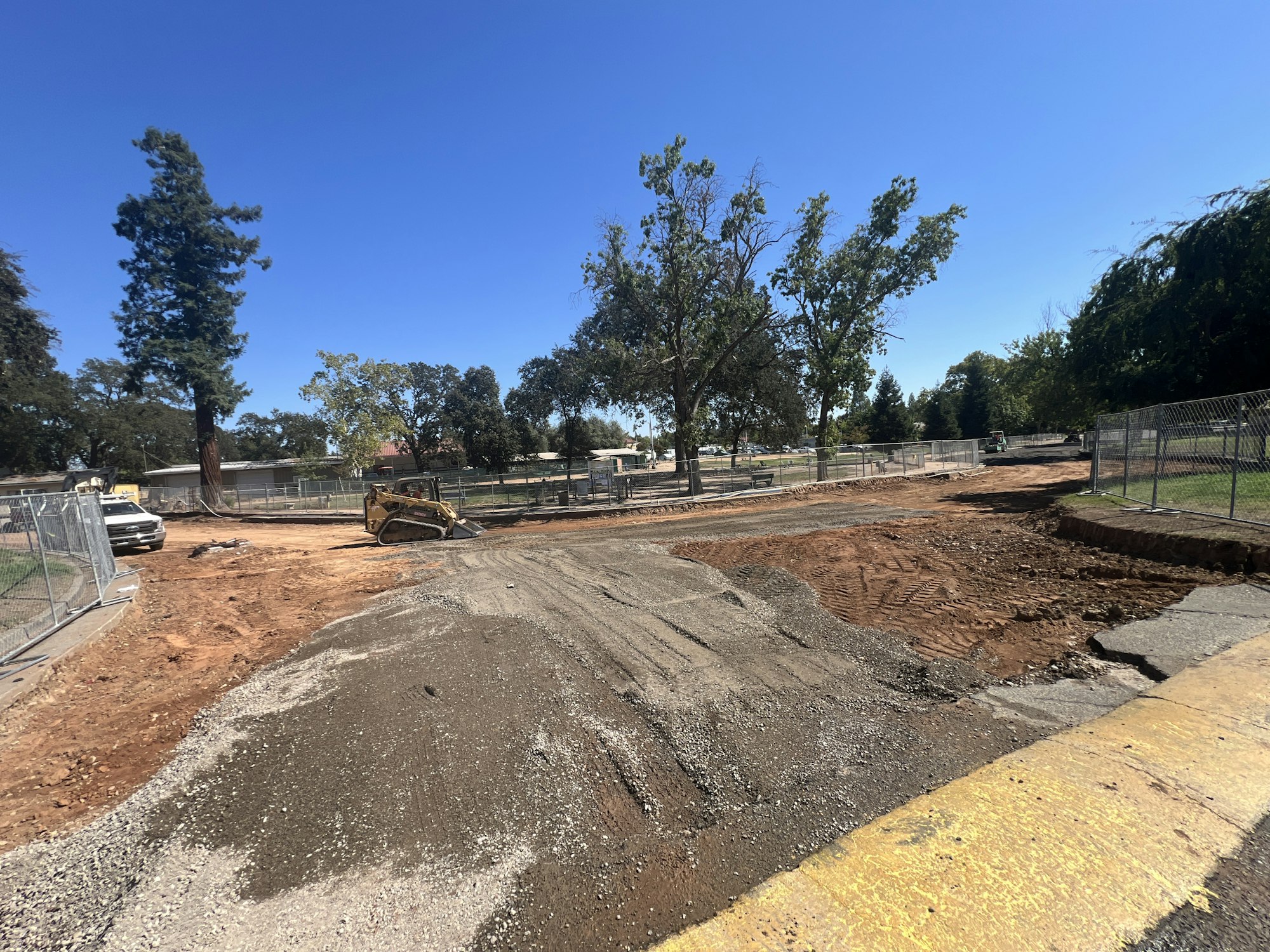 Construction site with a bulldozer, dirt, gravel, and trees in the background, fenced off with clear skies above.