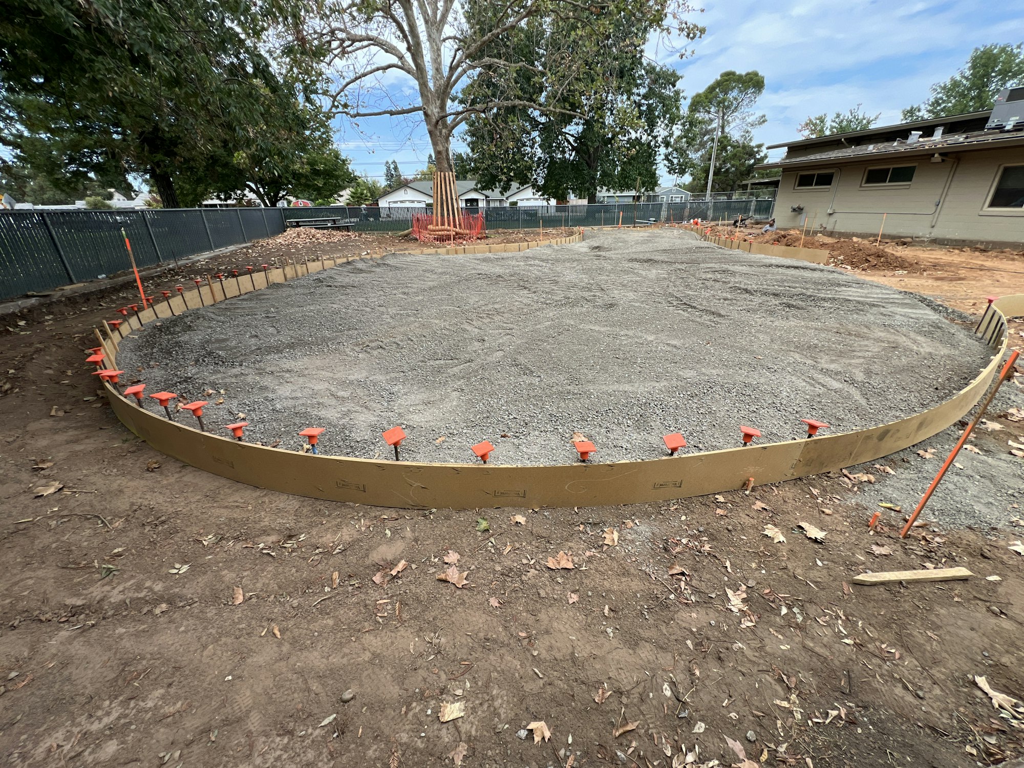 A circular construction area with gravel, enclosed by wooden forms and orange markers, near a building and trees.