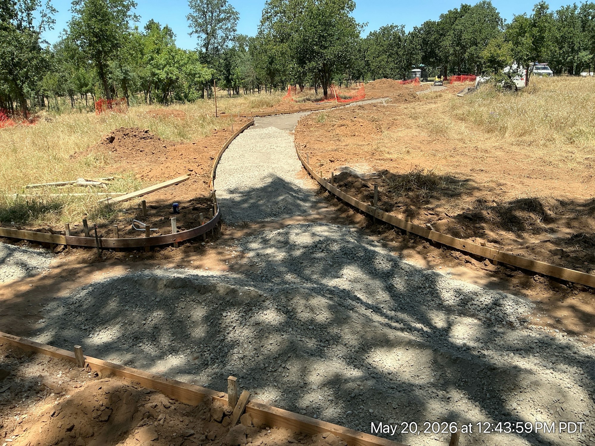 The image shows a construction site with a gravel path being shaped, framed with wooden boards, surrounded by grass and trees.