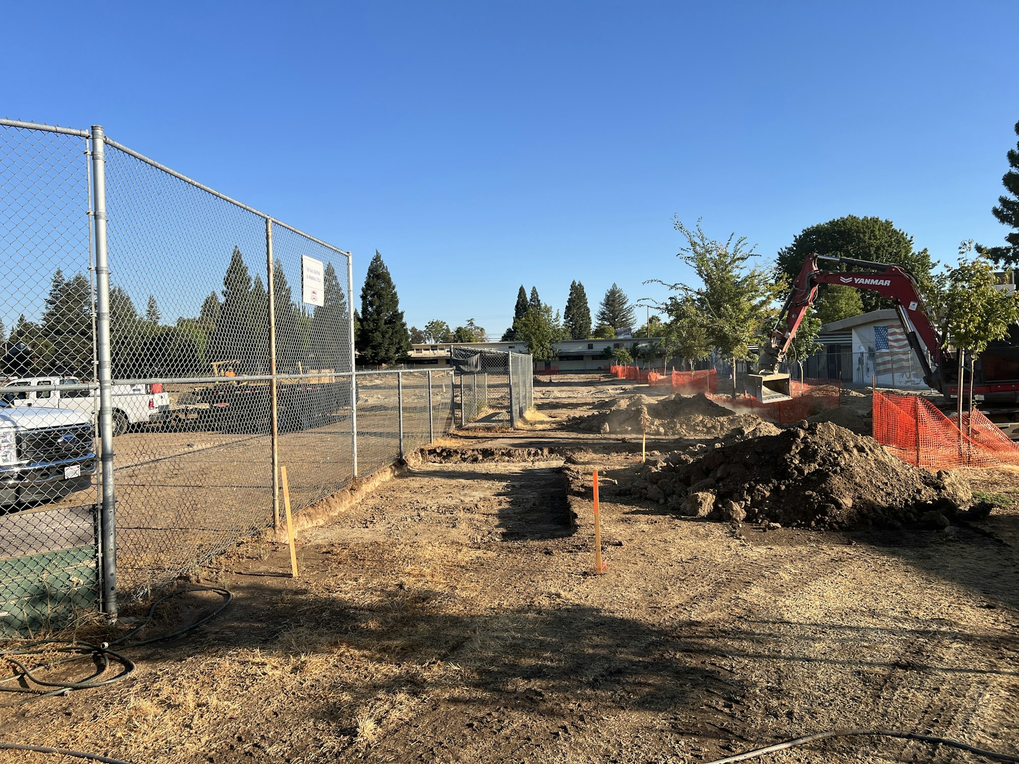 Construction site with excavator, fenced area, dirt piles, and trees in the background.
