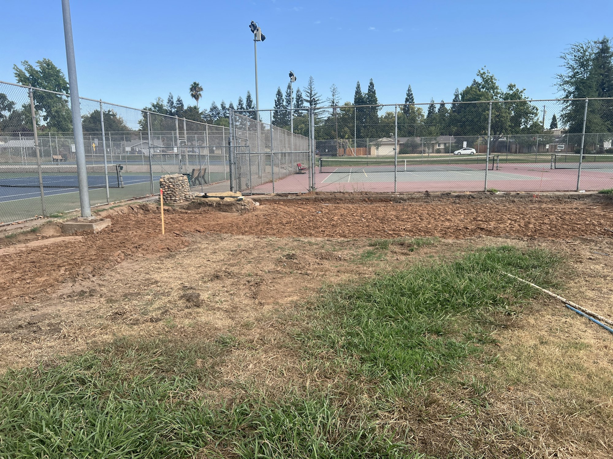 A fenced tennis court area with adjacent grassy and dirt patches, featuring lighting poles and a small pile of stones.