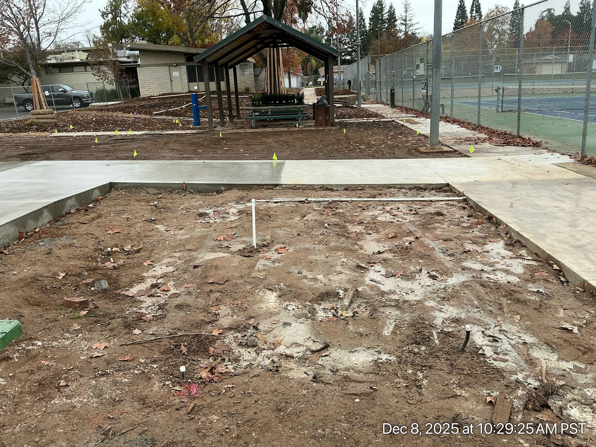 A park area under construction with partial concrete work, dirt, and yellow flags marking the site. Nearby structures include a shelter and tennis courts.