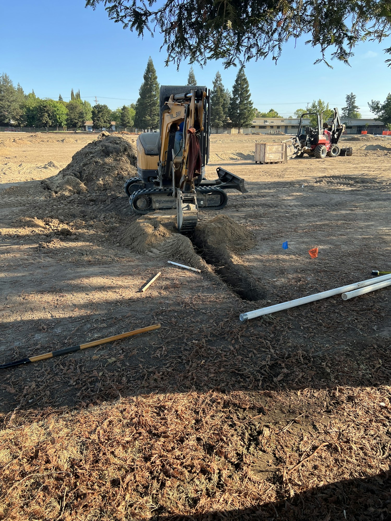 Construction site with a small excavator digging a trench, tools scattered, and trees in the background.
