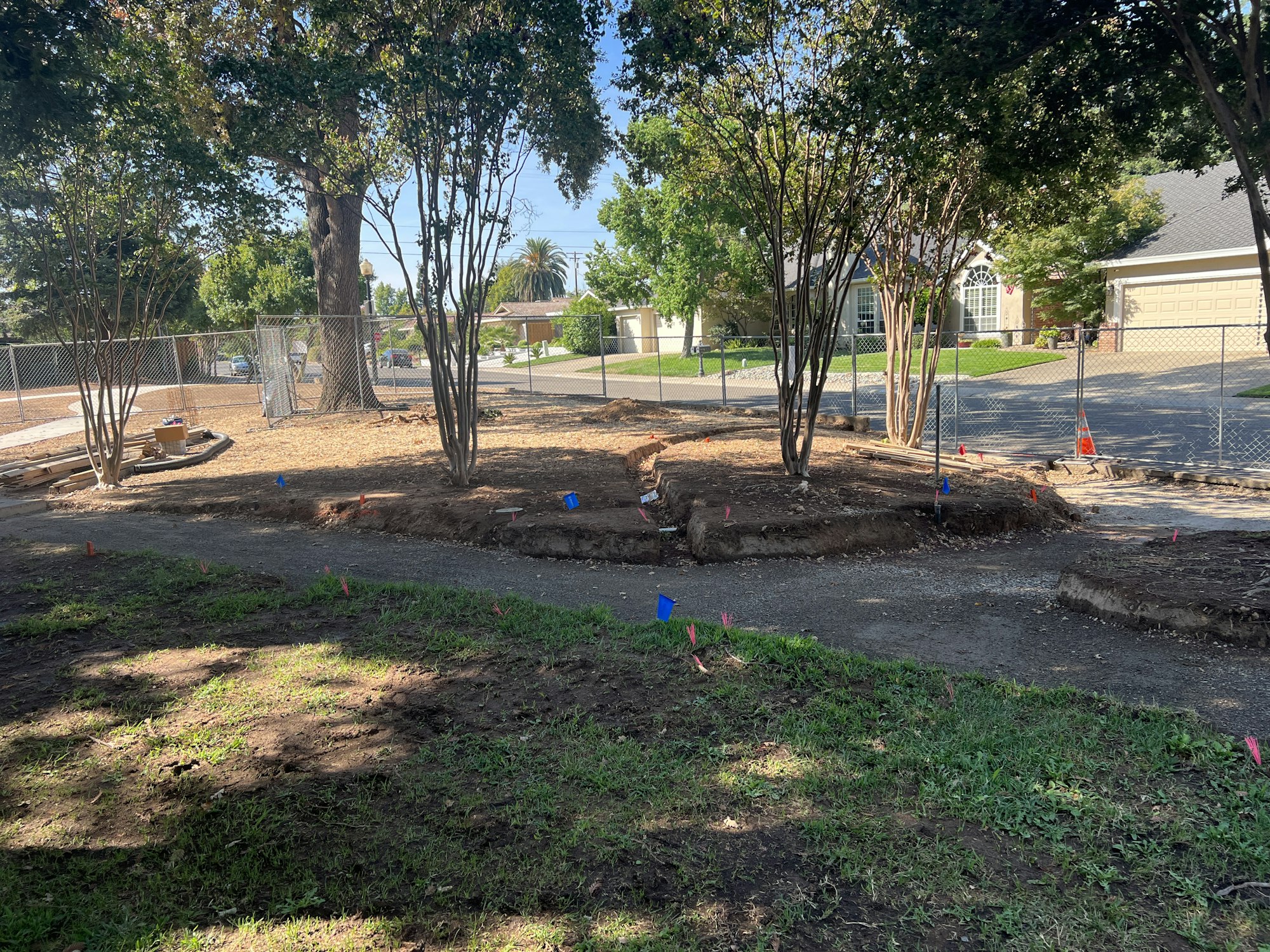 A fenced construction area with trees, dirt pathways, colored flags, and some materials.