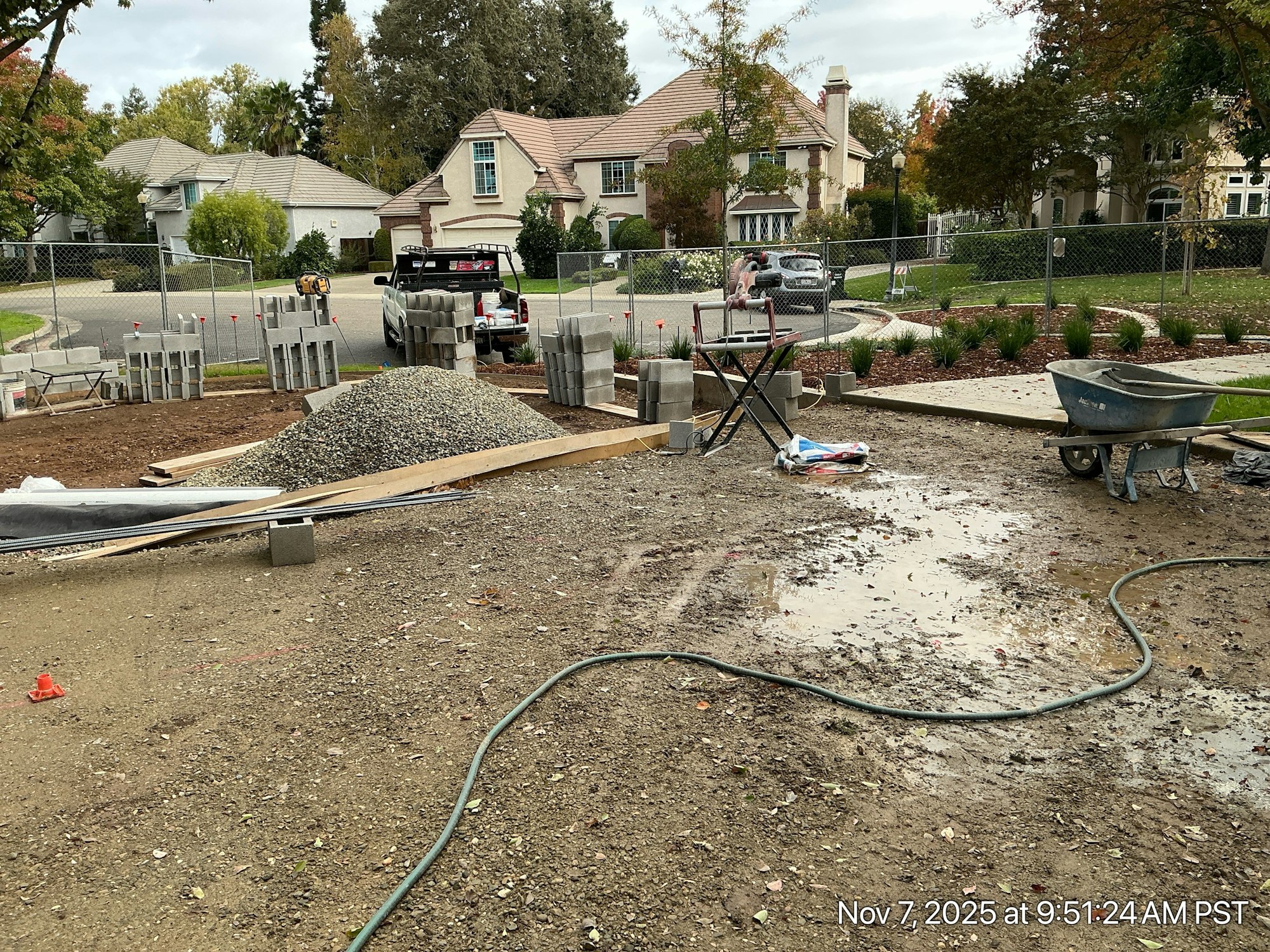 The image shows a construction site with gravel, concrete blocks, tools, and a wheelbarrow, set in a residential neighborhood.