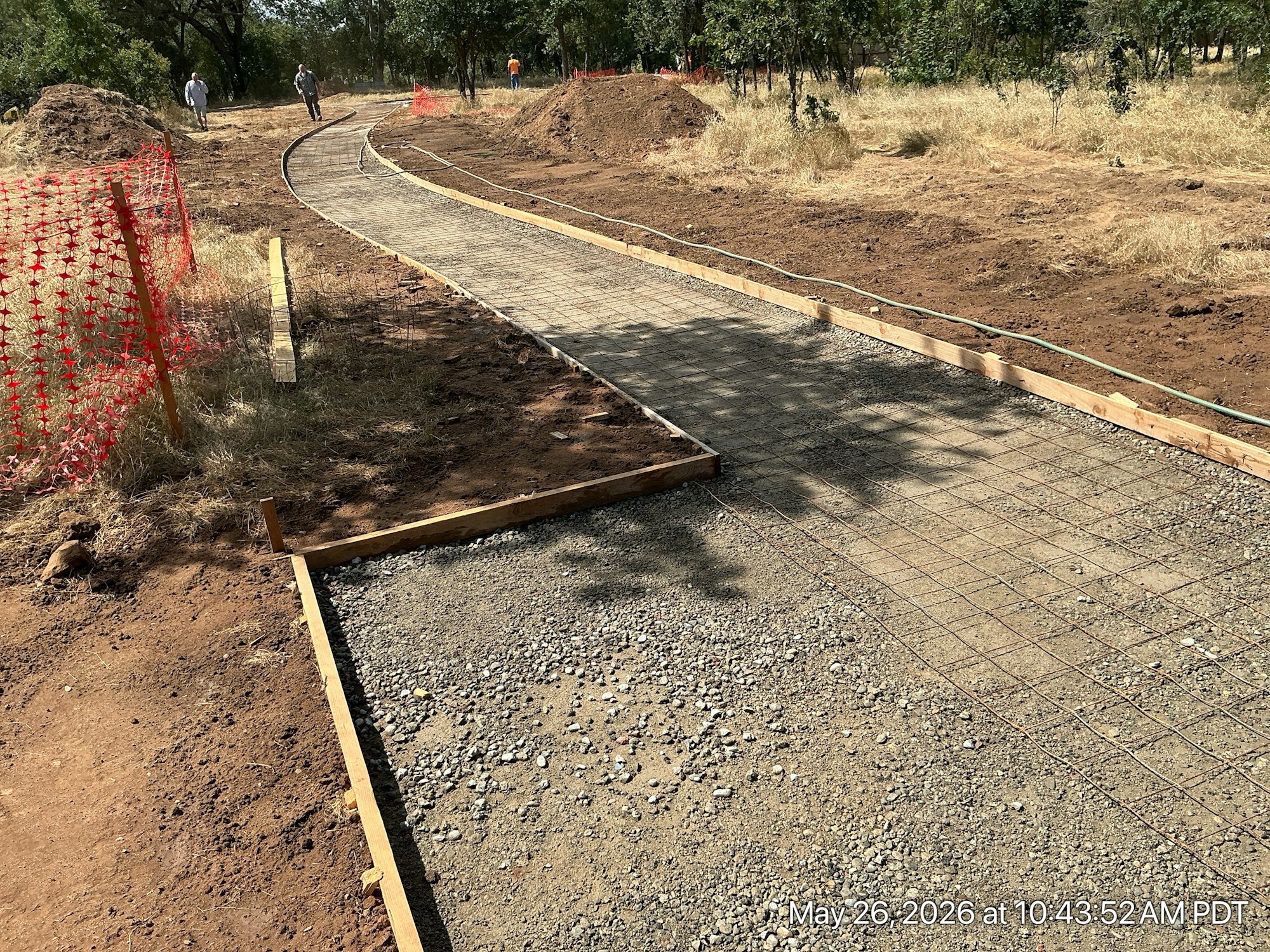 The image shows a paved pathway under construction, surrounded by dirt and red safety fencing, with workers in the background.