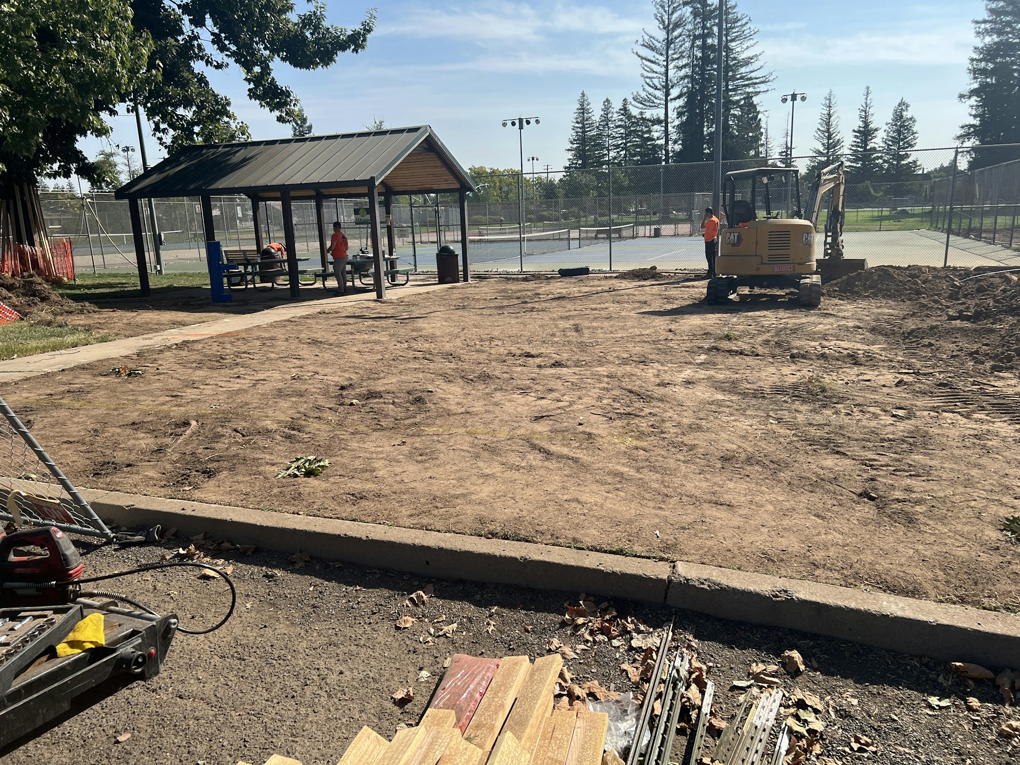 Construction site near tennis courts, with a small excavator, a pavilion, and equipment on the ground.