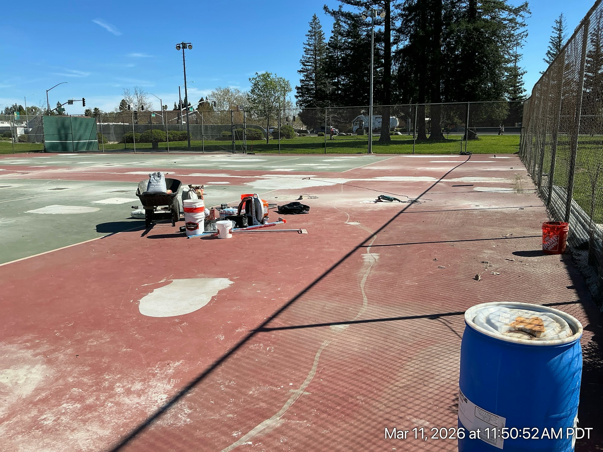 The image shows a partially worn-out tennis court with construction supplies and tools scattered around on a sunny day.