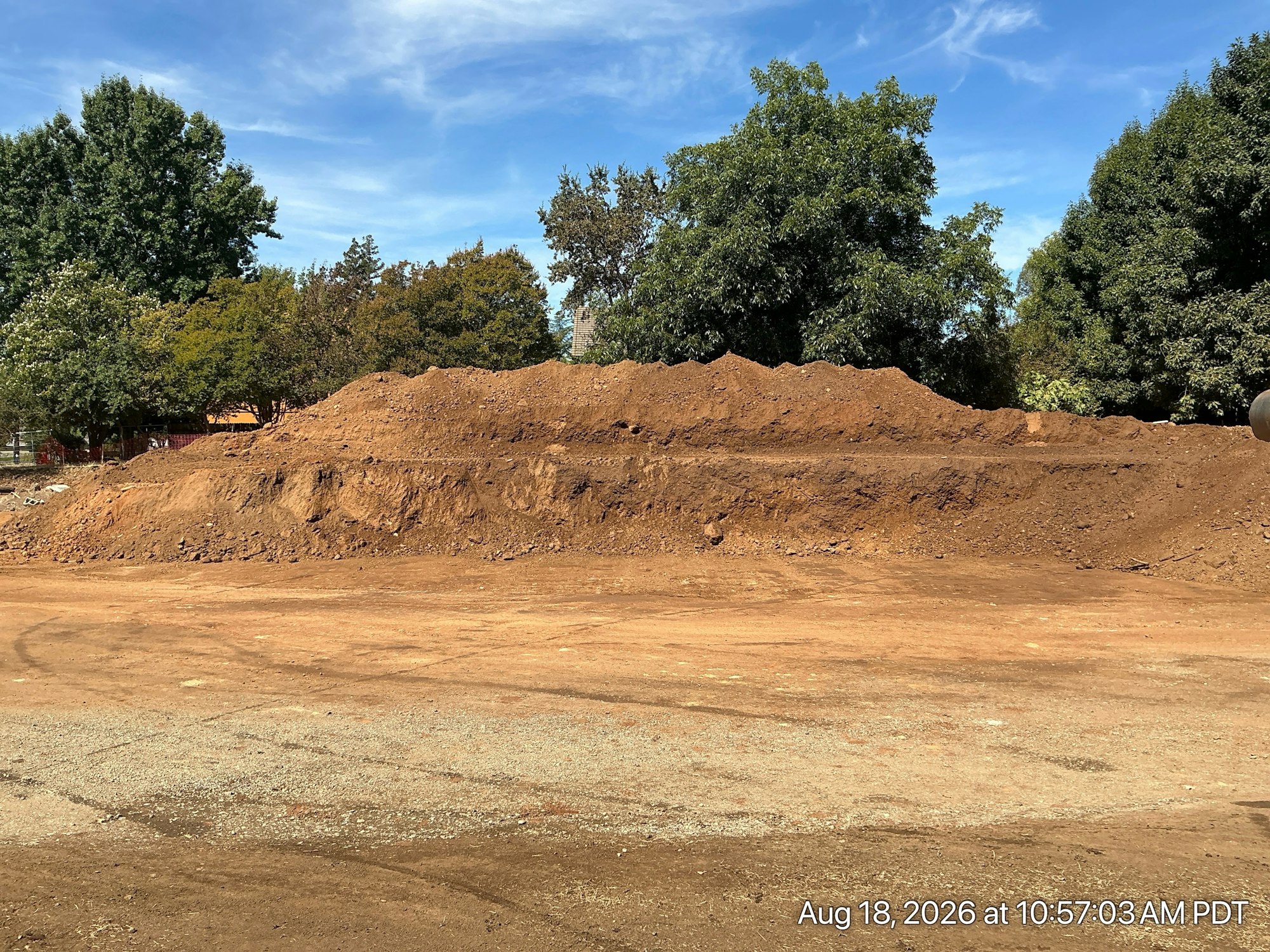 The image shows a large pile of dirt and gravel with trees in the background under a blue sky.