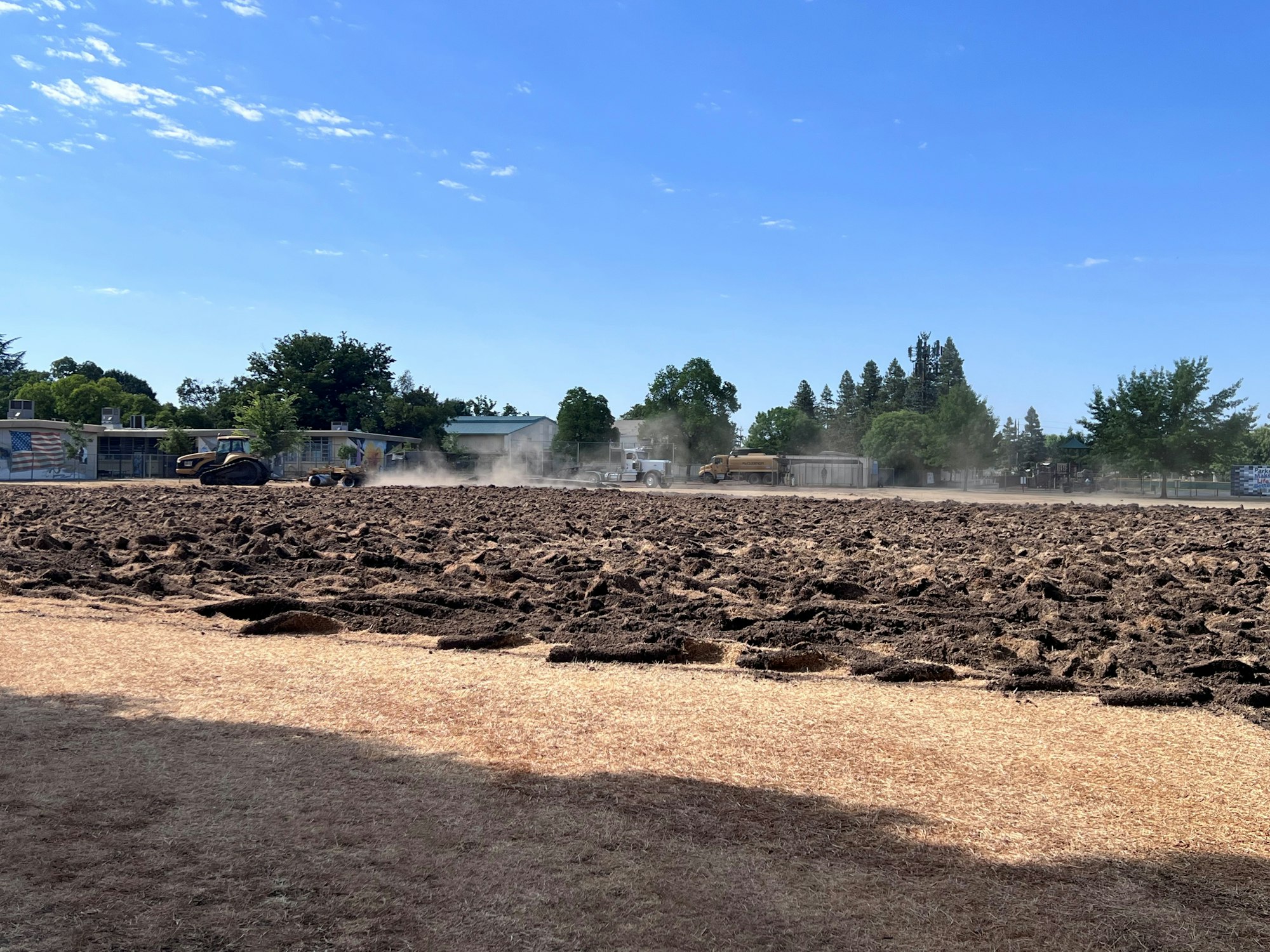 A field being plowed with machinery, with dust in the air and buildings and trees in the background under a clear blue sky.