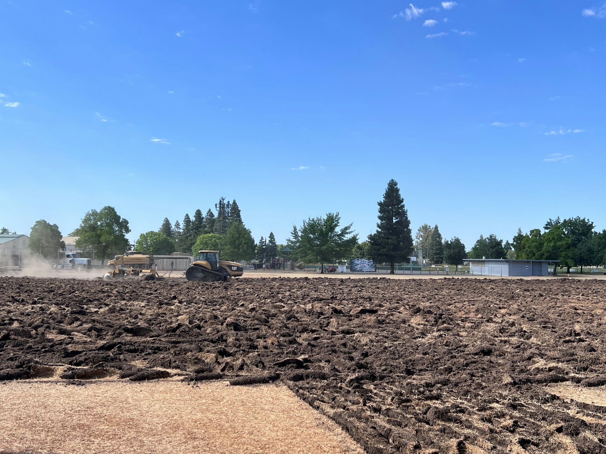 Construction vehicles working on a large dirt field with trees and buildings in the background under a clear blue sky.