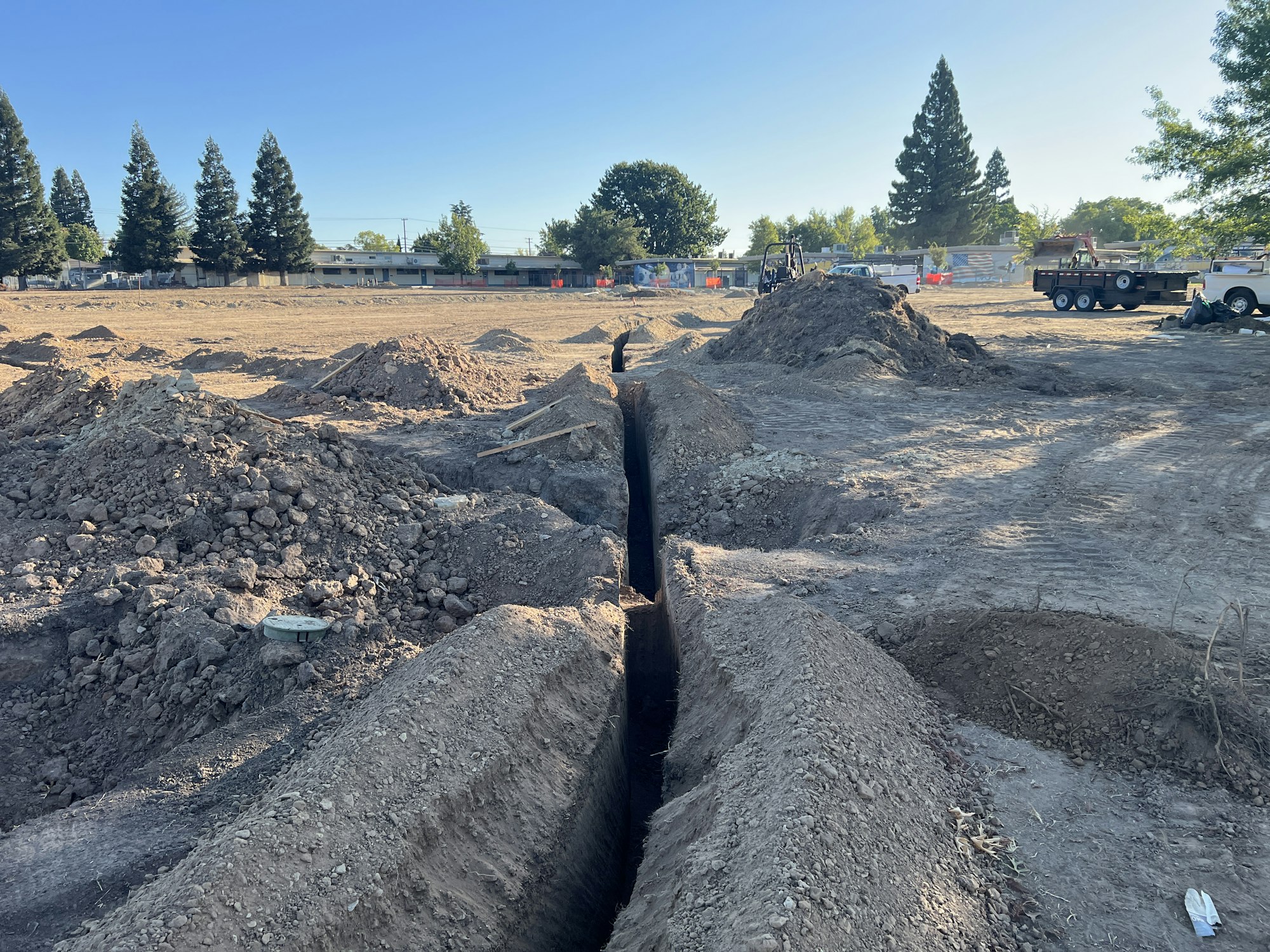 Construction site with dug trenches, dirt piles, and parked vehicles under a clear sky.