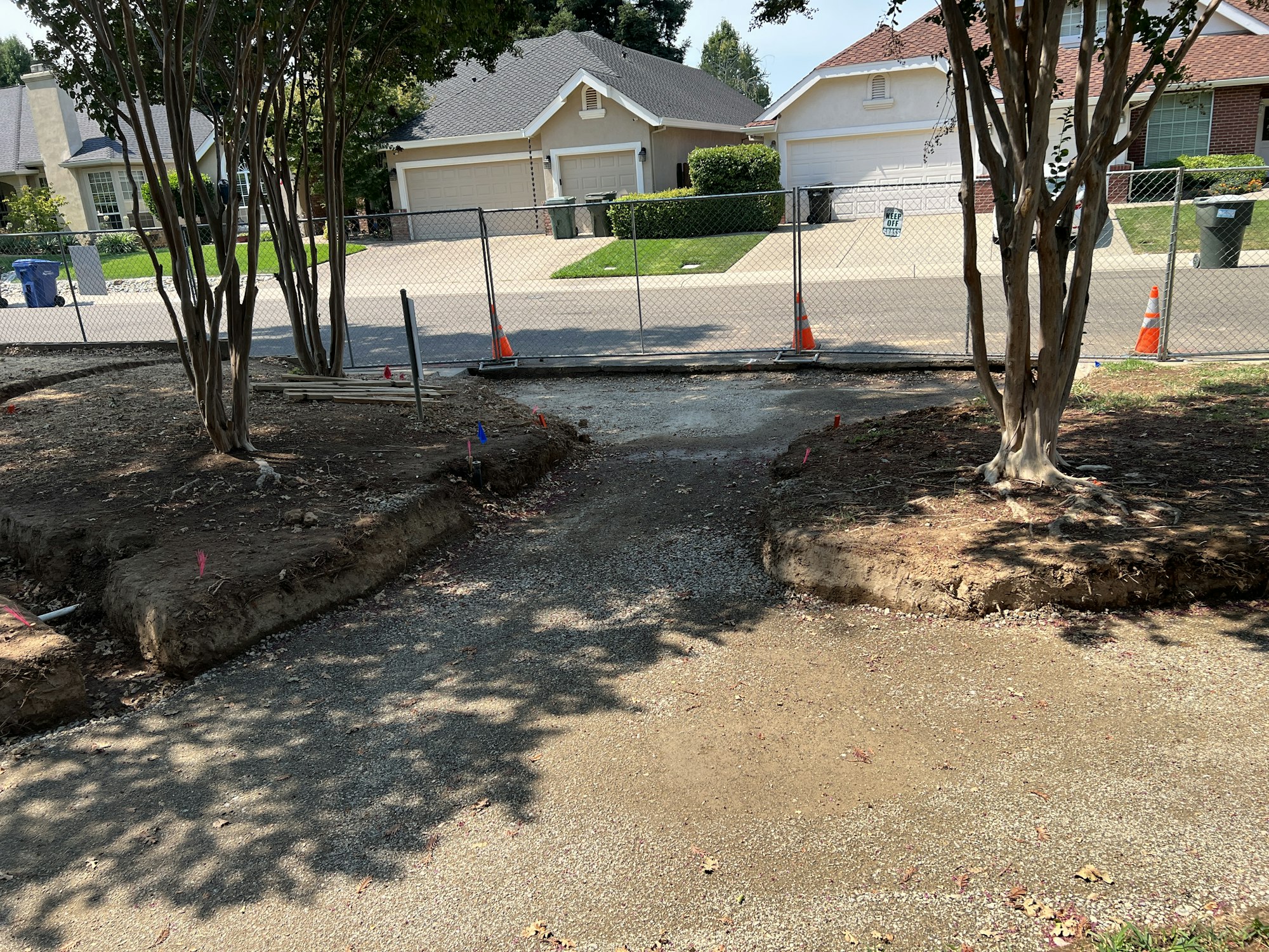 A construction area with trees, dirt, safety cones, and a fenced-off section in a residential neighborhood.