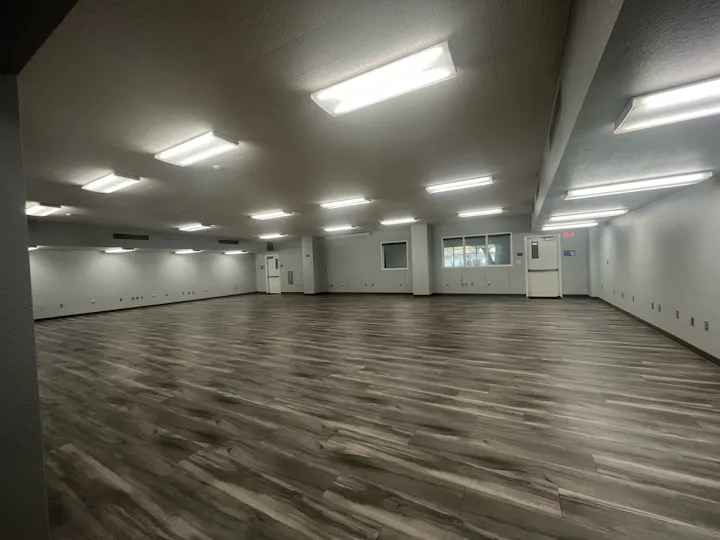 The image shows a spacious, empty room with gray walls, large lighting, and wood-patterned flooring.