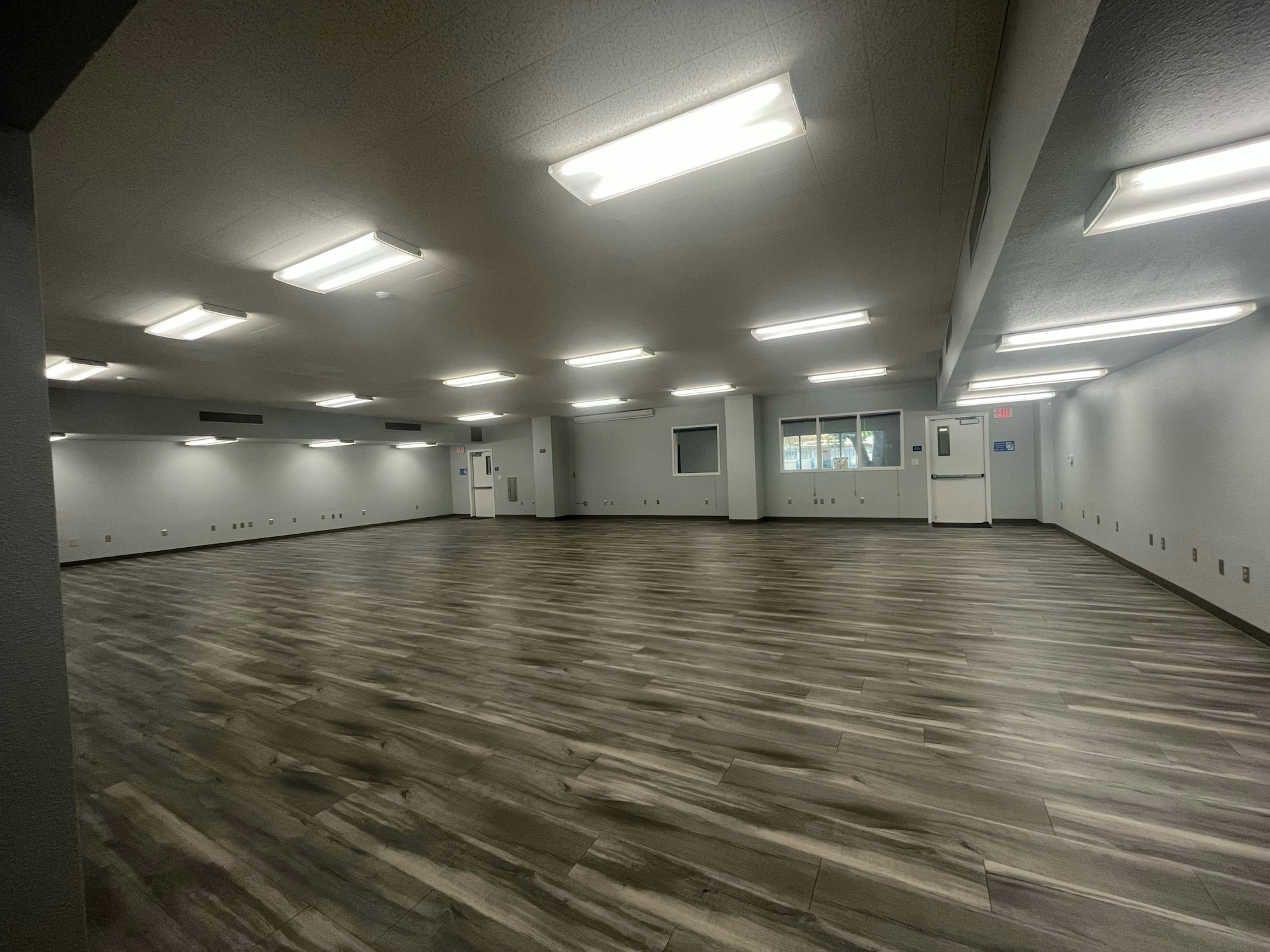 The image shows a spacious, empty room with gray walls, large lighting, and wood-patterned flooring.