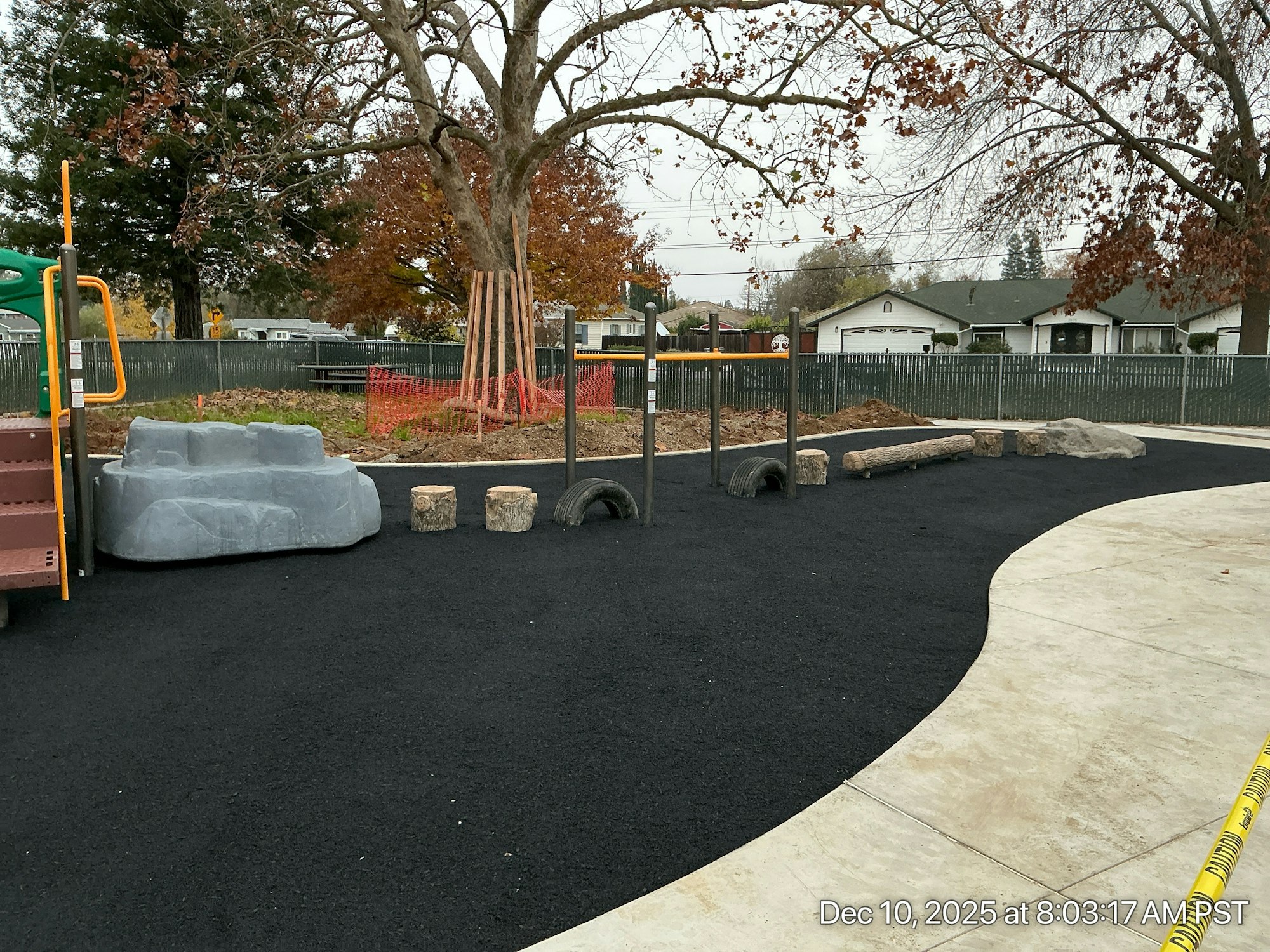 A playground area featuring climbing structures, logs, rocks, and a rubberized surface, surrounded by a fence and trees.