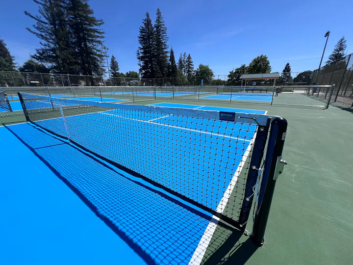 An outdoor tennis court with vibrant blue surfaces, nets, and surrounding trees, under a clear blue sky.