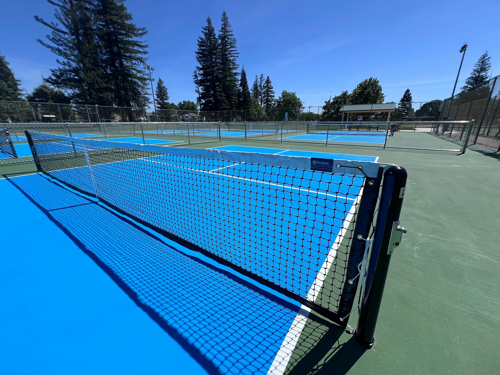 An outdoor tennis court with vibrant blue surfaces, nets, and surrounding trees, under a clear blue sky.