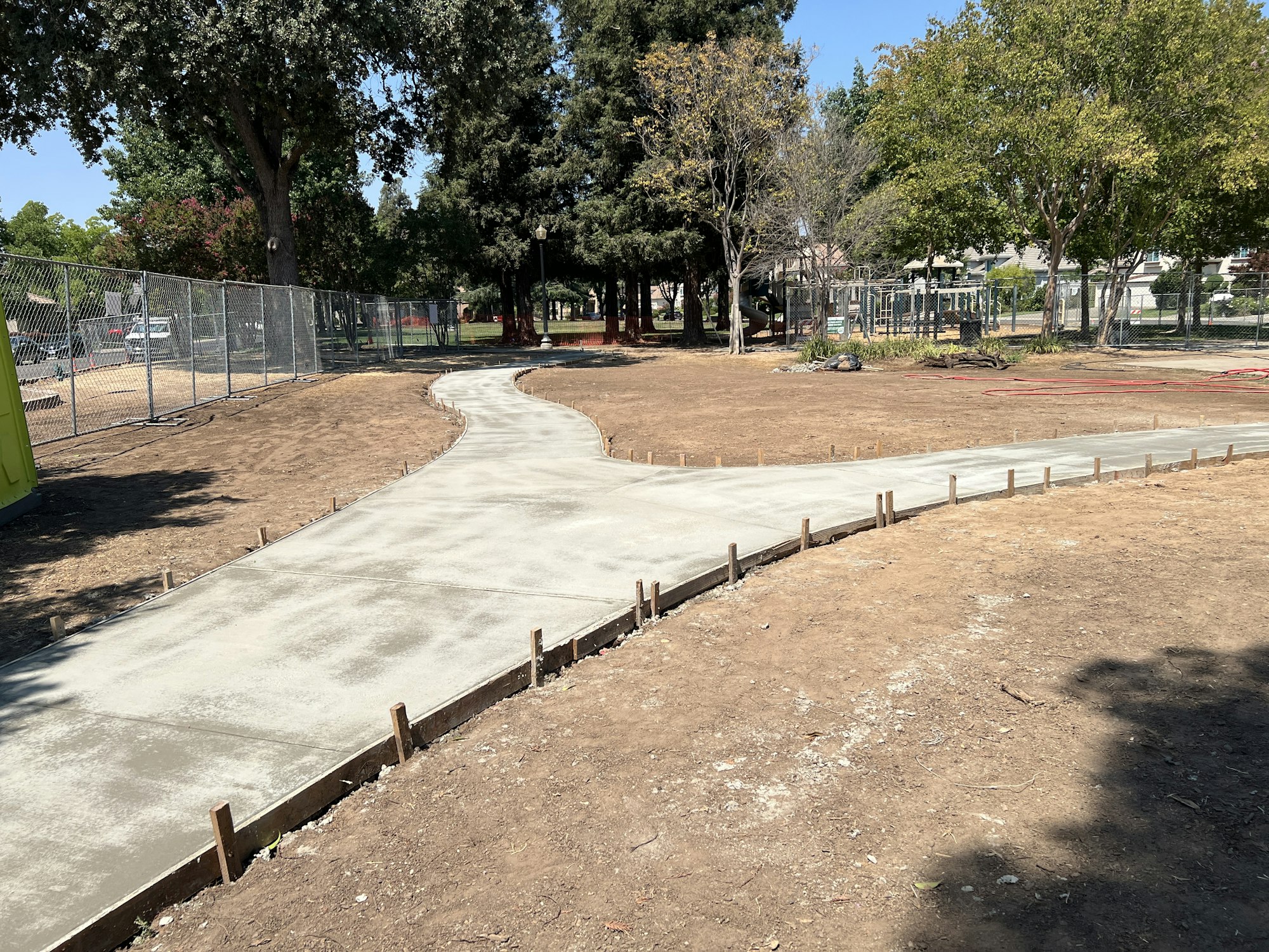 A freshly paved concrete path curving through a park area with trees and a construction fence.