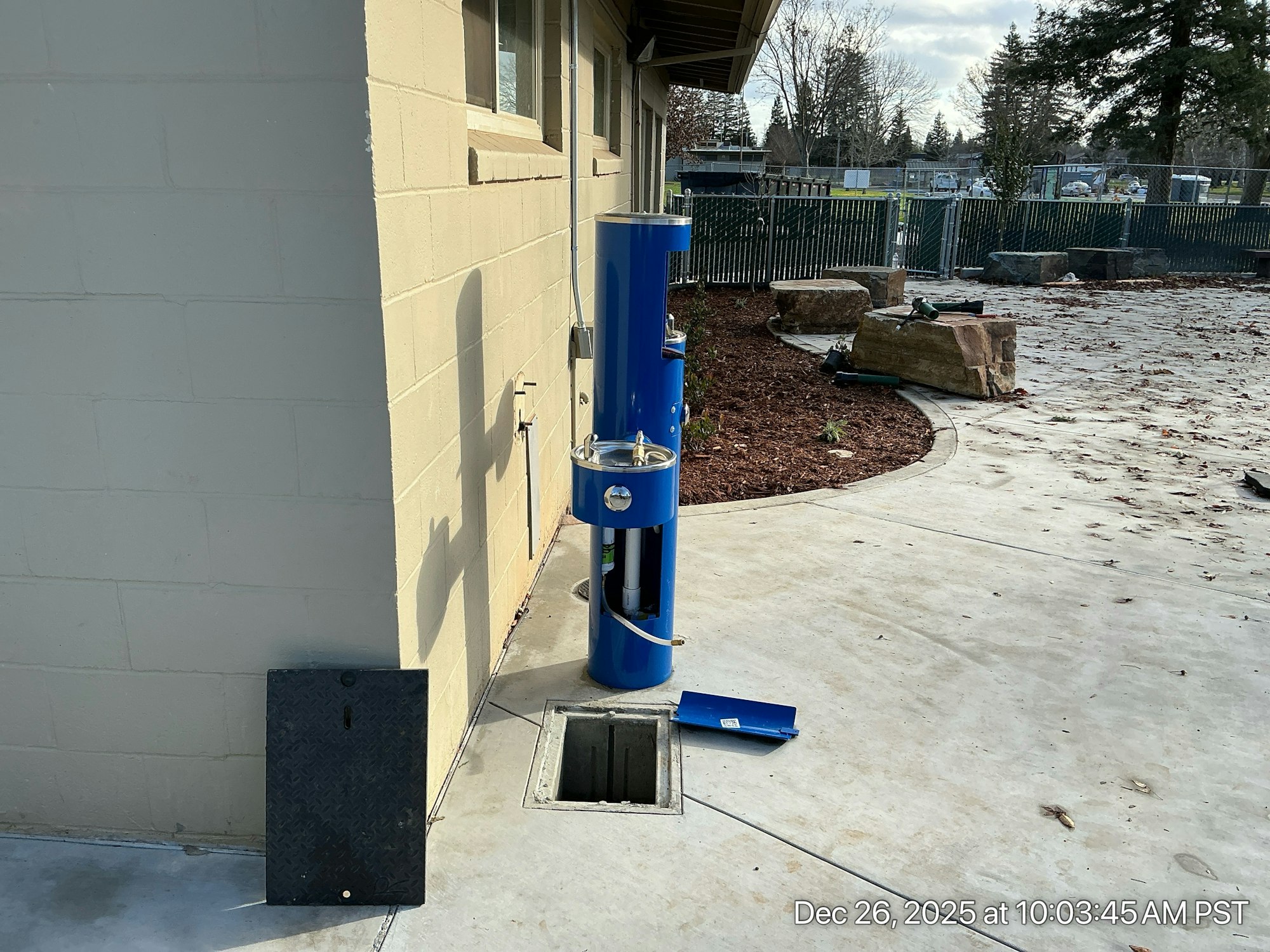 An outdoor blue structure, likely a drinking fountain, next to a wall, with a removable cover on the ground nearby.